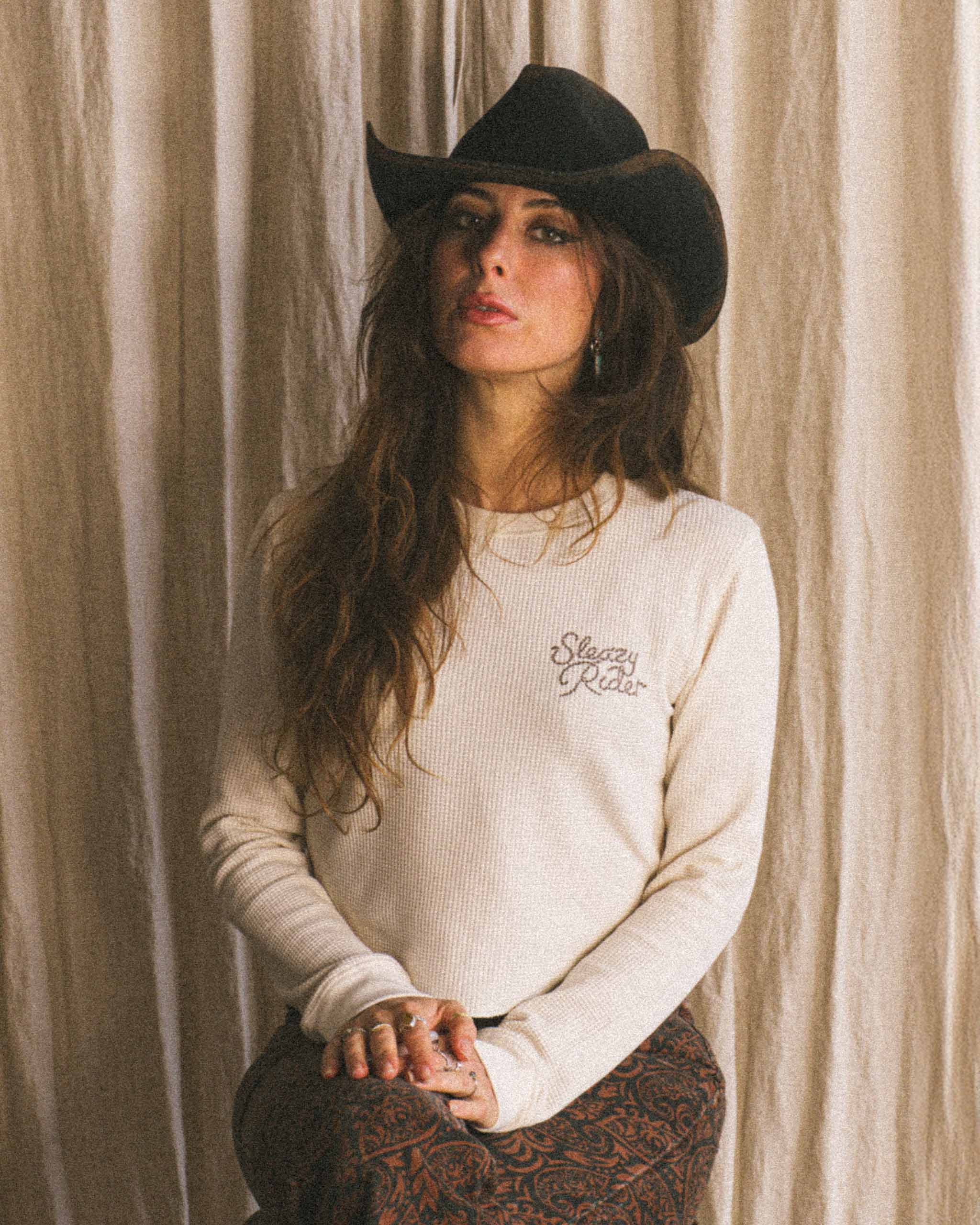 A cowgirl wearing a white waffle knit long sleeve with Sleazy Rider printed on it.
