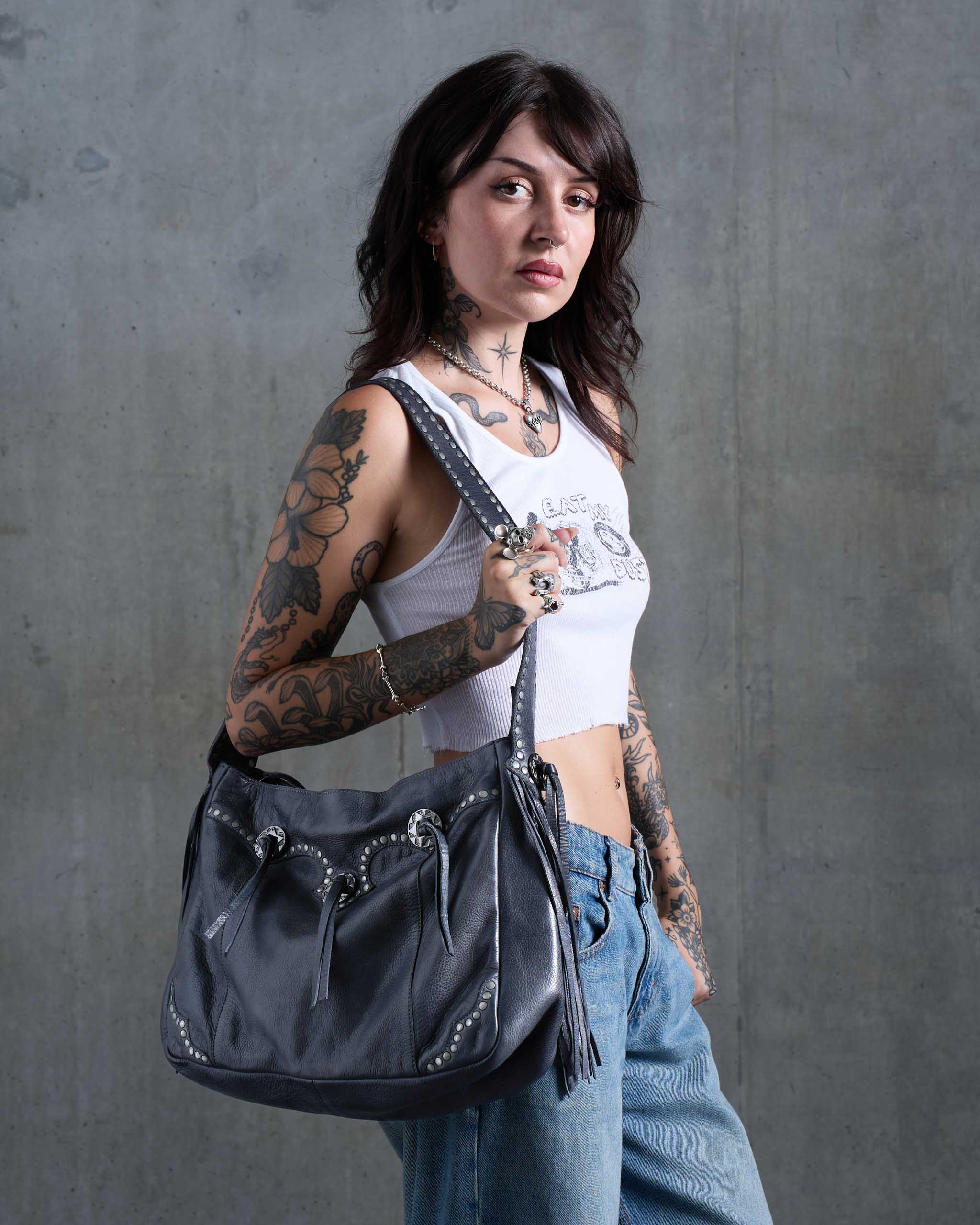 A tattooed woman carries a grey western leather shoulder bag with silver studs and tassels against a gray concrete wall.