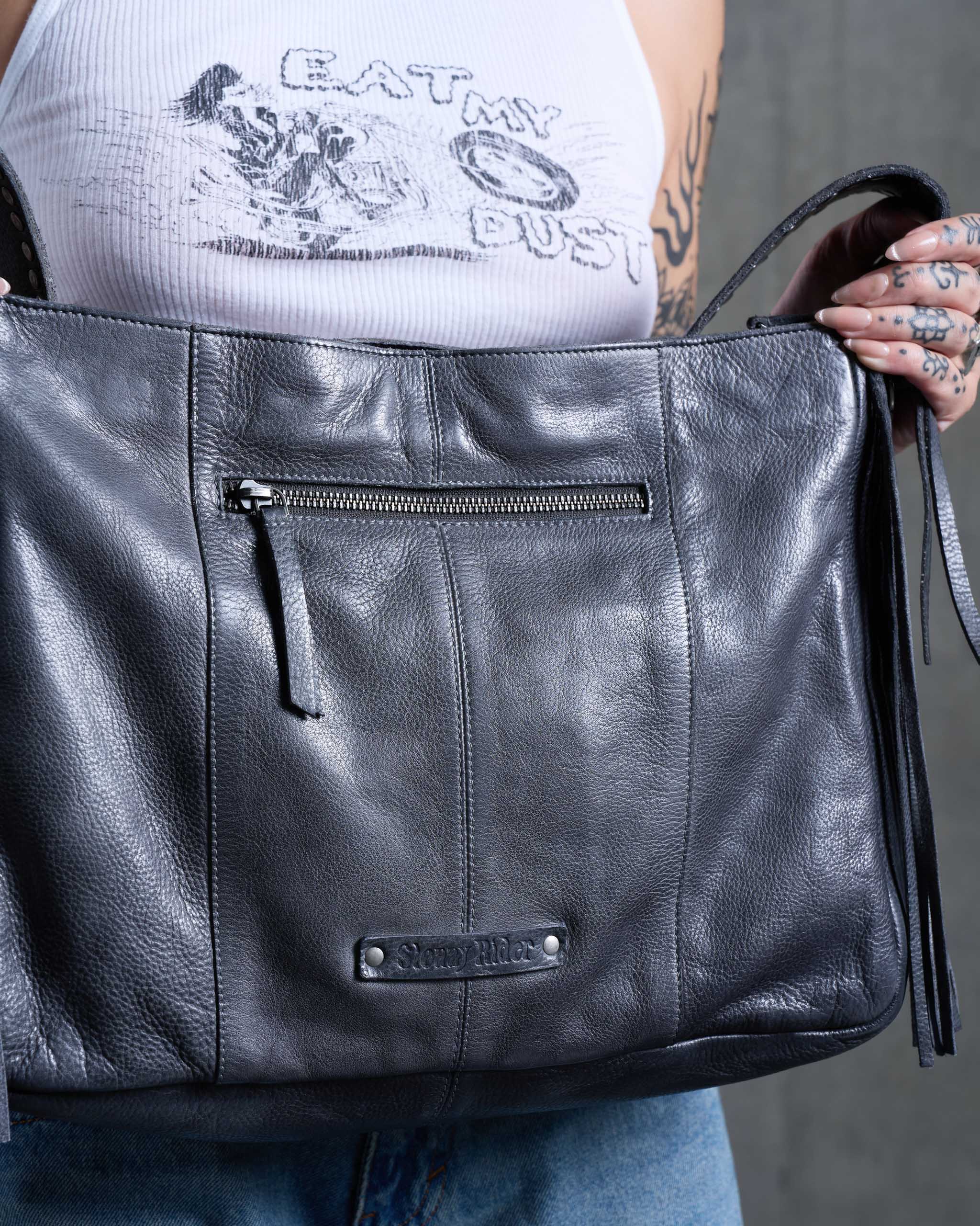 A tattooed woman holding a dark gray western leather bag, with a 'Sleazy Rider' leather tag.