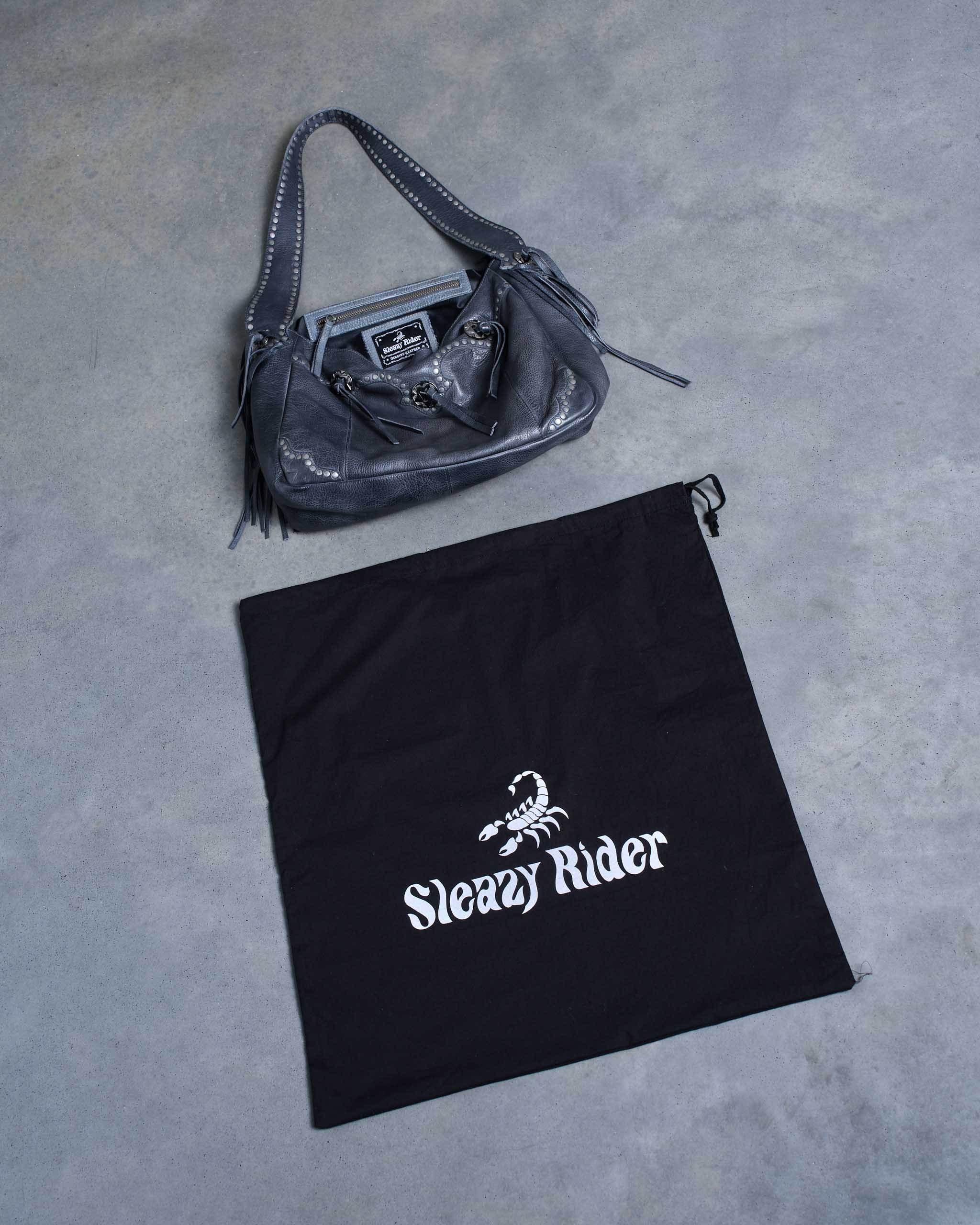 A dark grey western bag, with studs, conchos and fringe; laid next to a black Sleazy Rider dustbag on a concrete floor.
