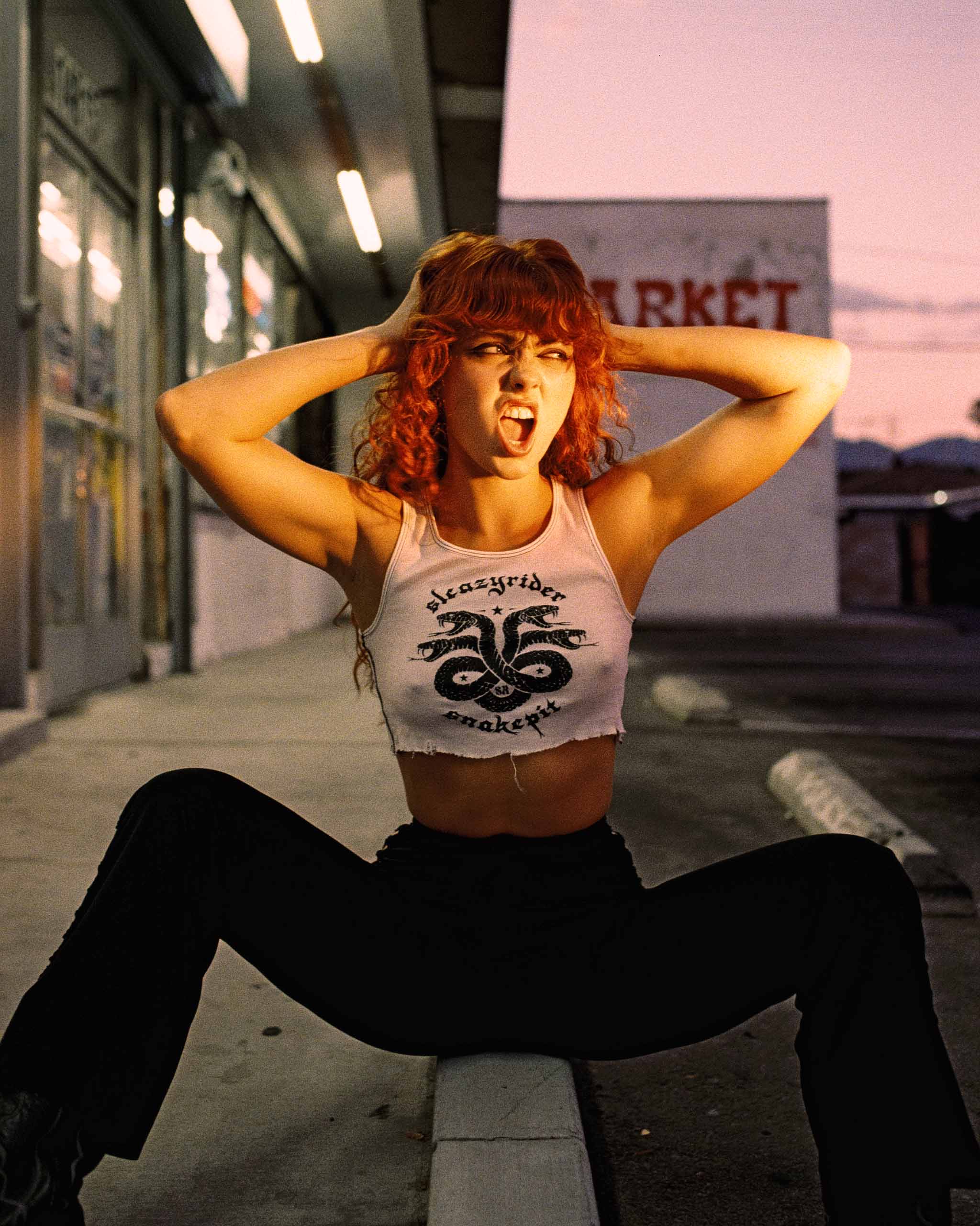 A redhead model wearing a white ribbed tank top with snake graphic sat roadside by a liquor store.