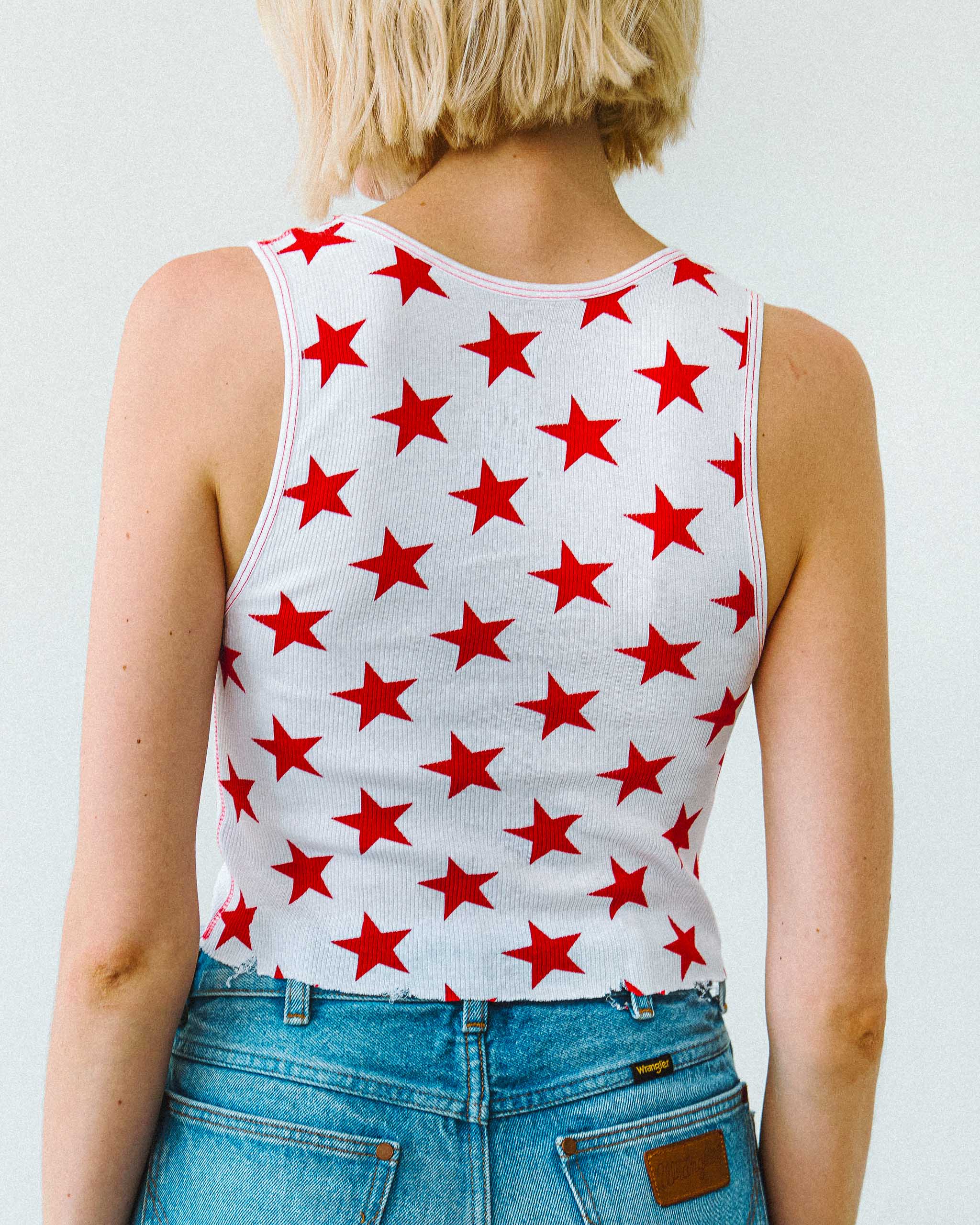 Model wearing a white tank top with red stars printed all over, and denim shorts on a plain background.