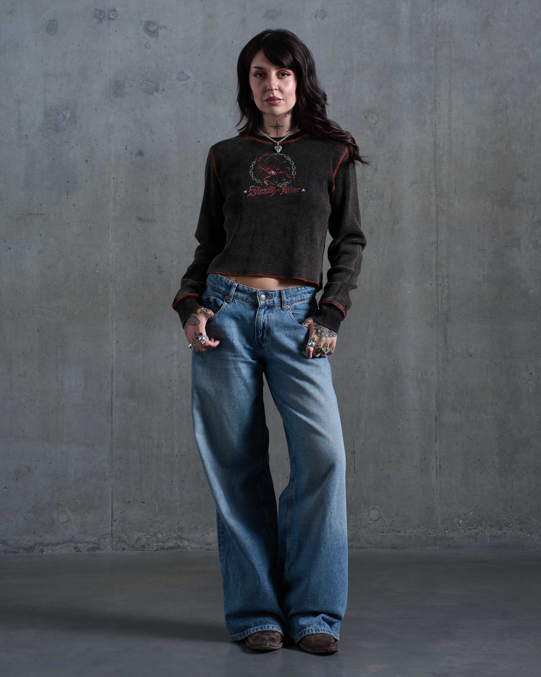 A biker woman wearing a dark waffle knit top with orange contrast stitching and a scorpion print, with low rise jeans and brown cowboy boots.