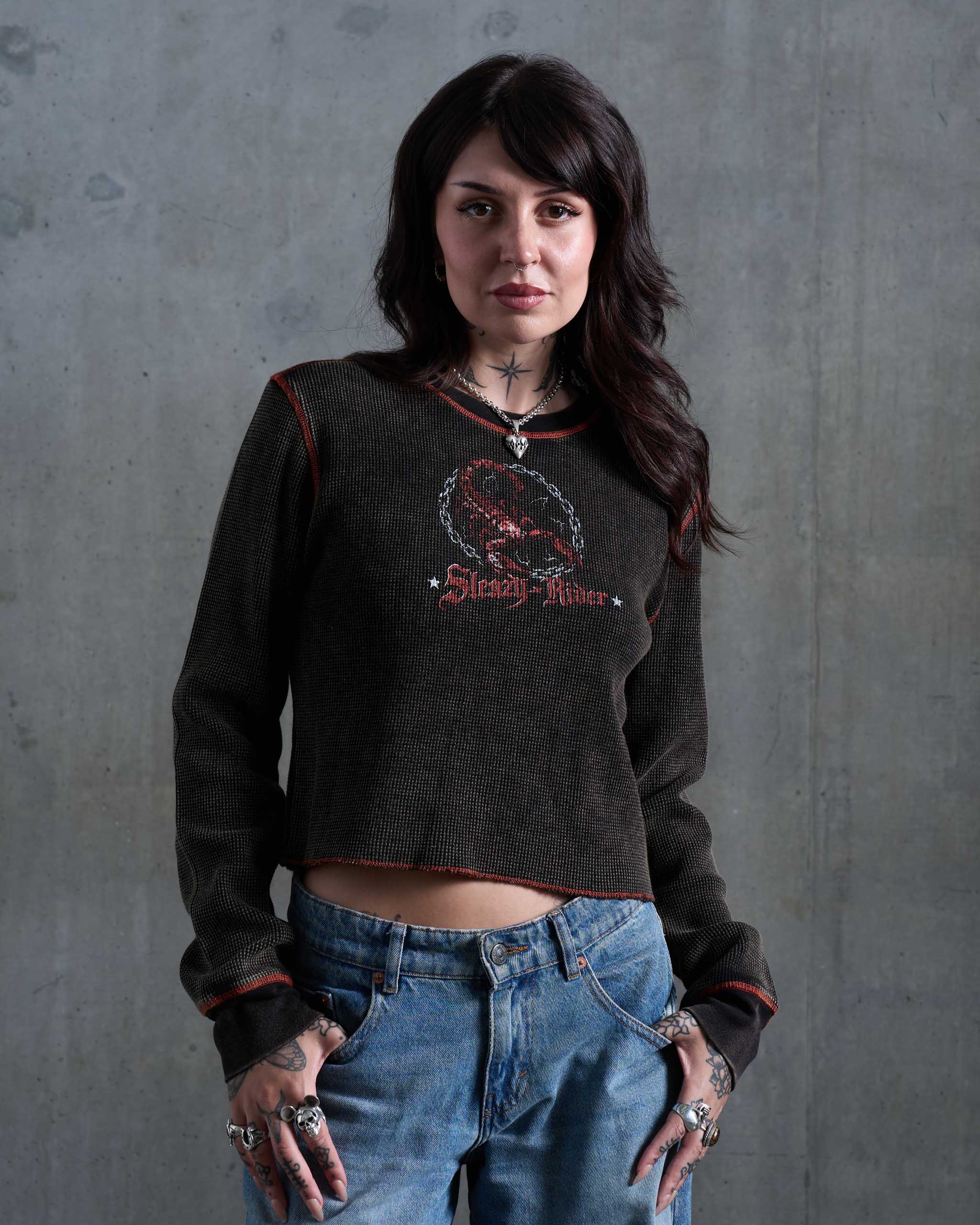 A biker woman wearing a dark waffle knit top with orange contrast stitching and a scorpion print, and low rise jeans.