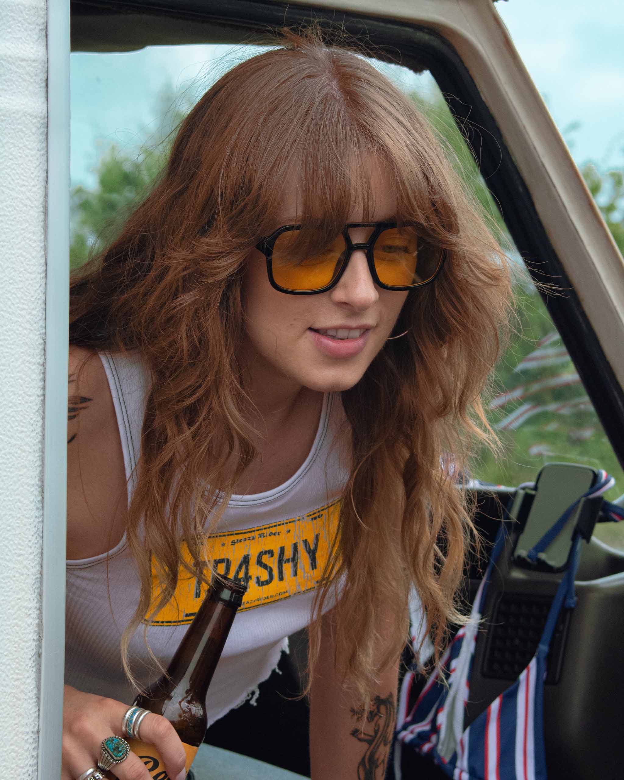 Woman with 70s sunglasses and a 'TRASHY' sign yellow and white tank top in a vintage camper van.