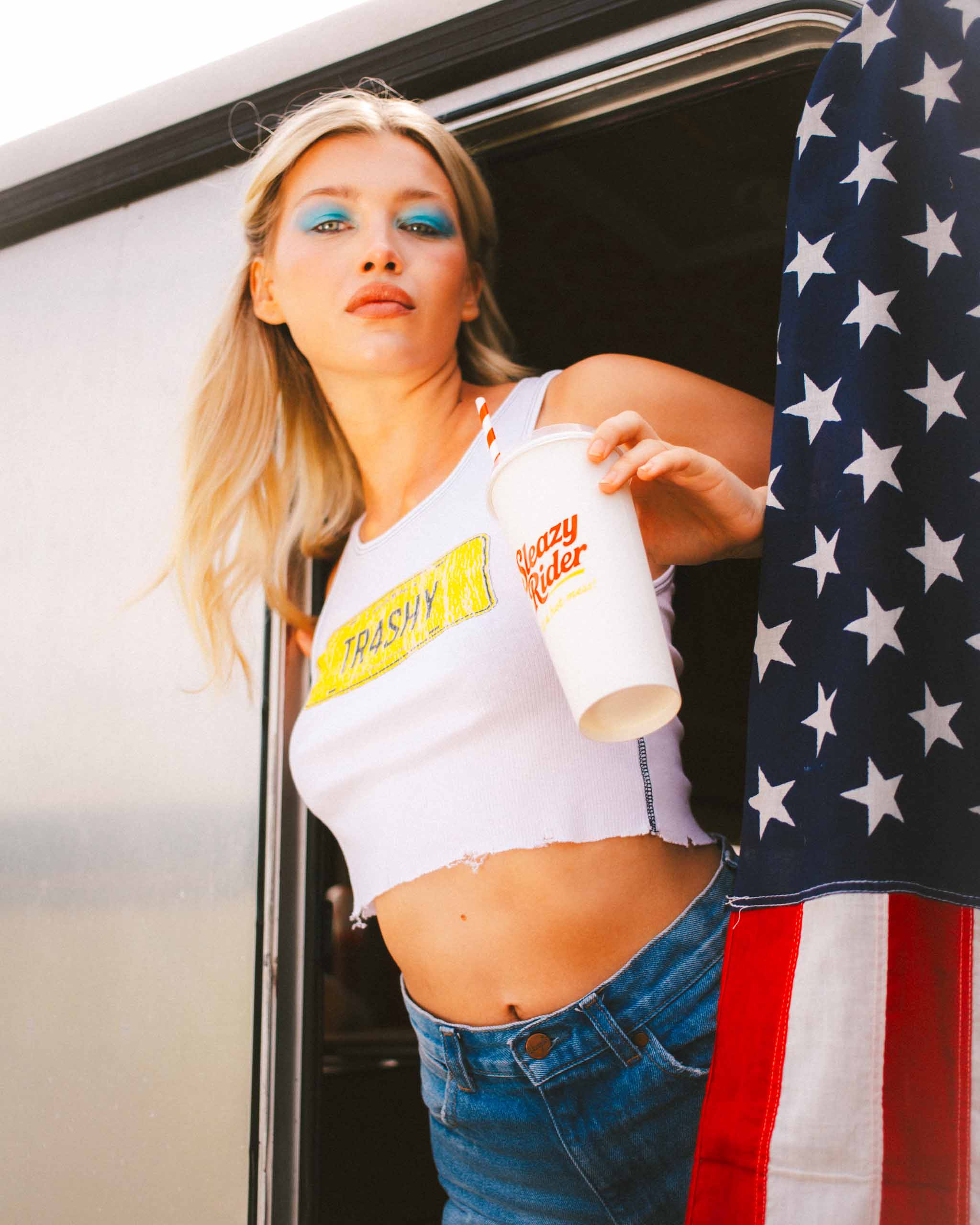Woman wearing a white tank top with black stitching with a "TR4SHY" graphic, holding a Sleazy Rider cup with an American flag in the background