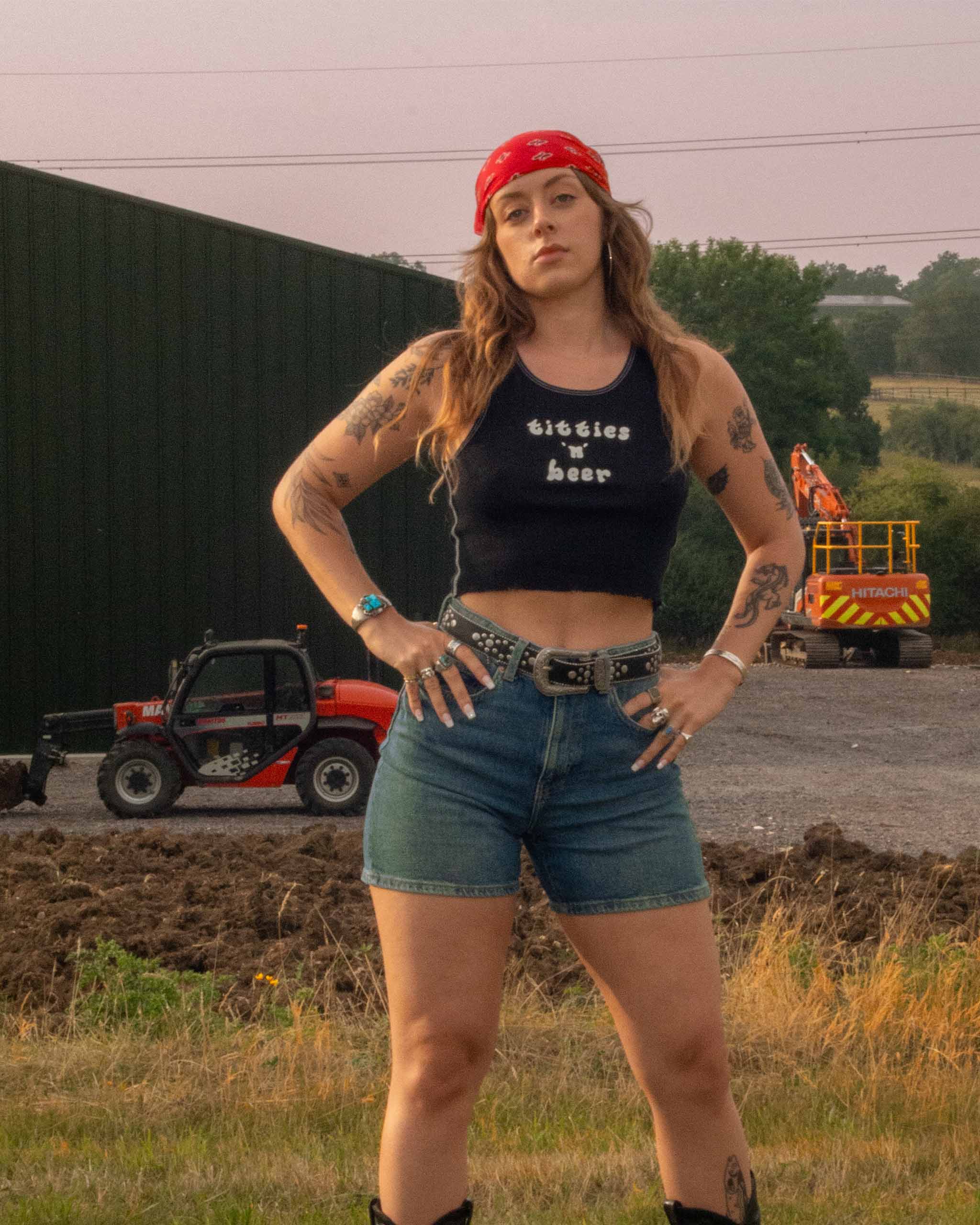 Woman wearing a black tank top with "Titties n Beer" text and denim shorts standing outdoors with tractors in the background.