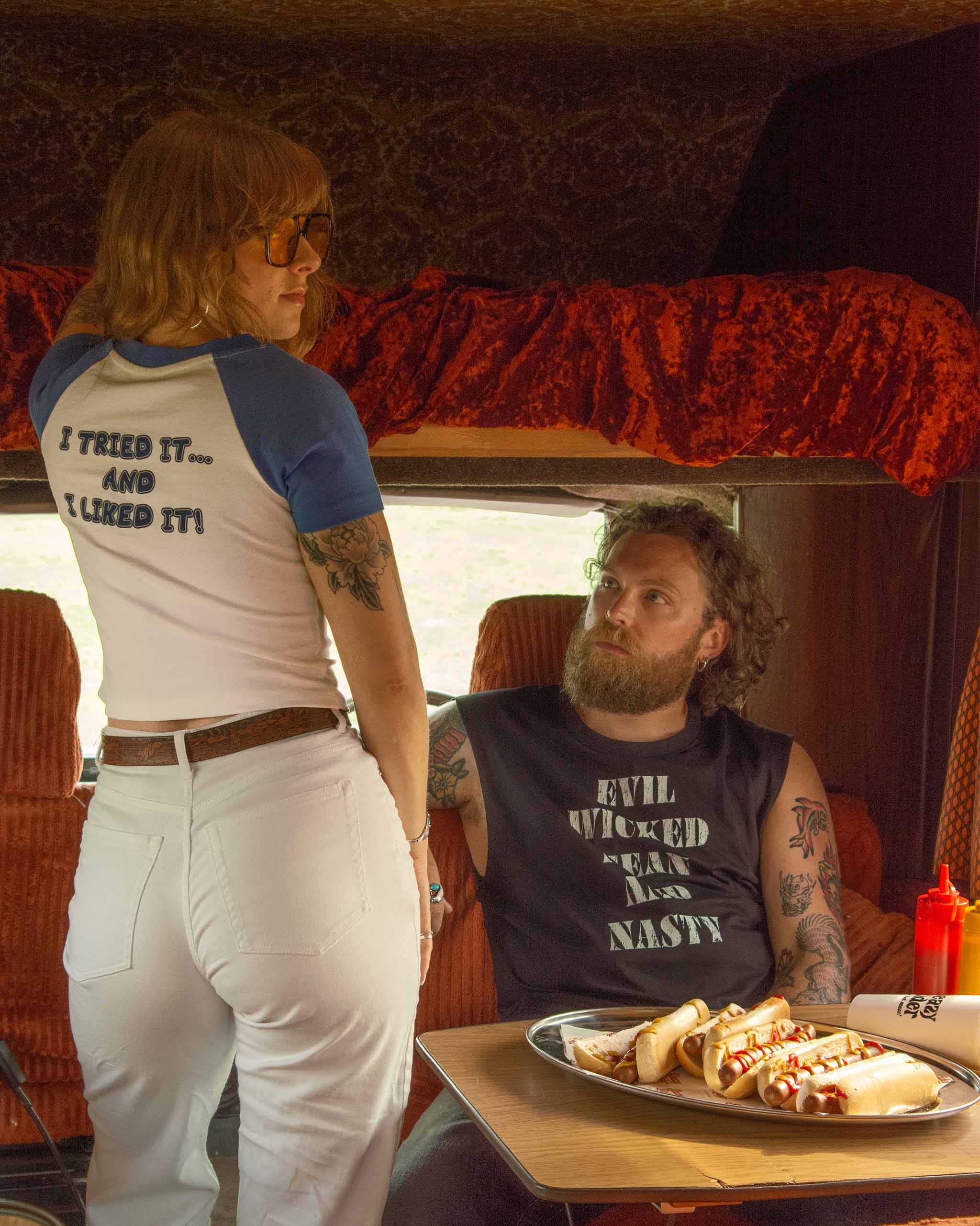 Two people inside a 70s camper with one person standing and the other sitting at a table with food. Woman is wearing a blue and white raglan baby t-shirt and man is in a muscle tank.