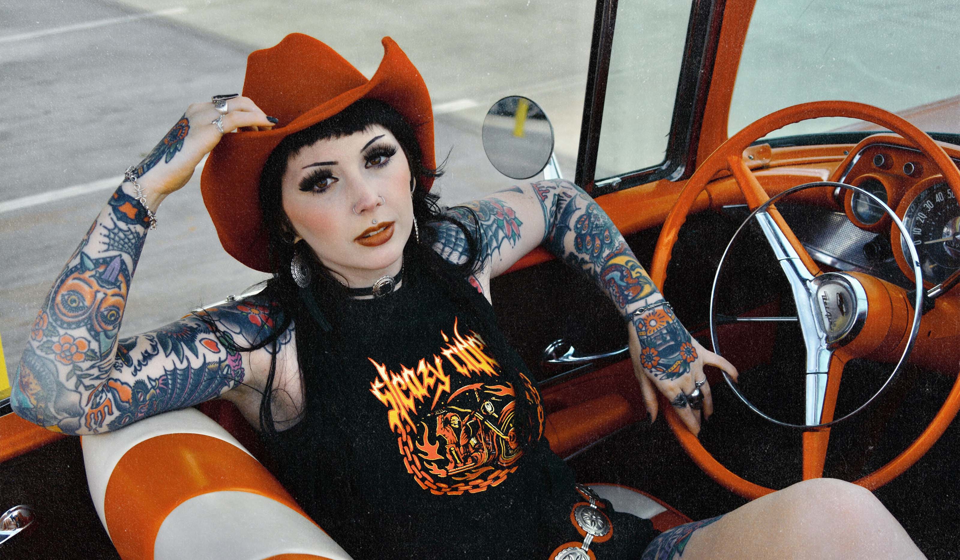 An alternative model in a vintage car wearing a red cowboy hat and a Sleazy Rider tank top