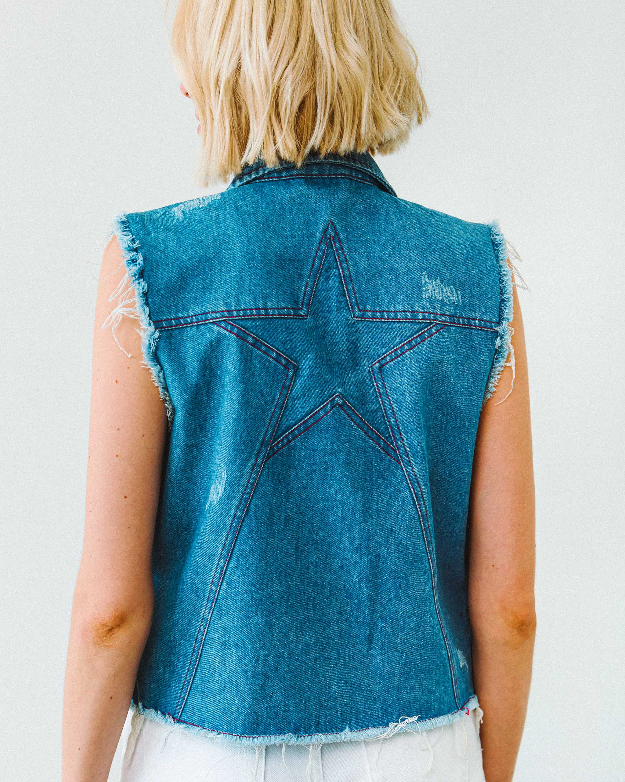 A woman wearing a denim cut off vest with a star panel on the back and vintage distressed detailing.
