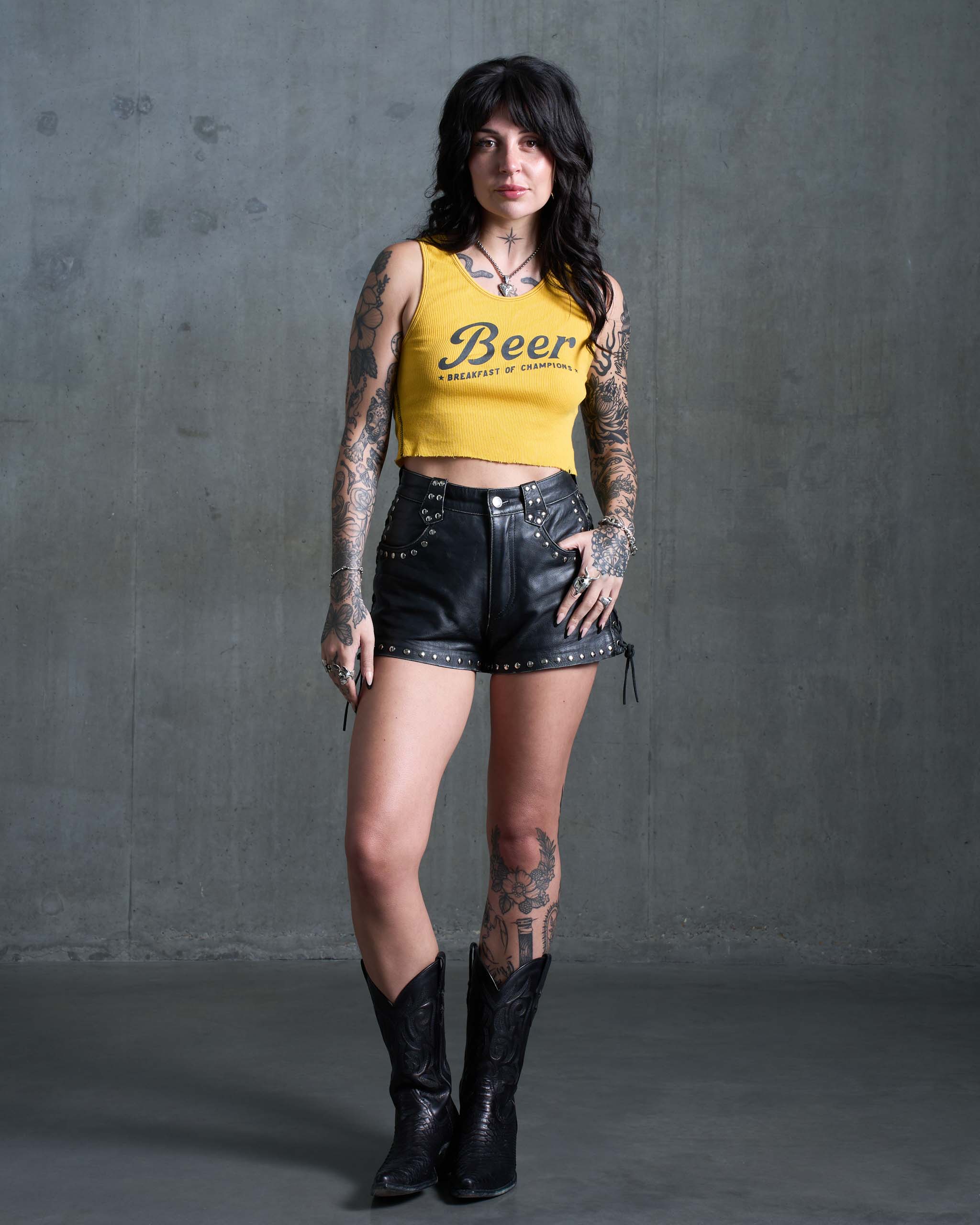 A tattooed woman wearing a yellow 'Beer' tank top, with studded leather shorts and black cowboy boots.