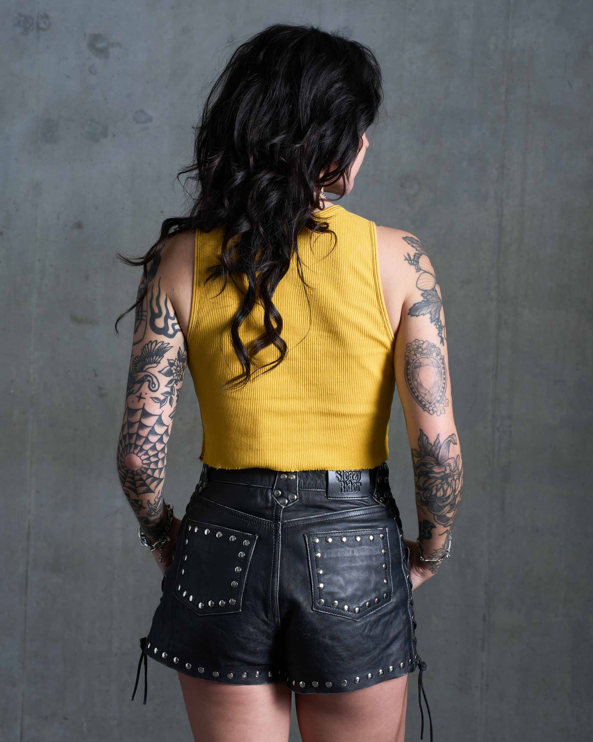 A tattooed woman wearing a yellow tank top and studded leather shorts, view from behind.
