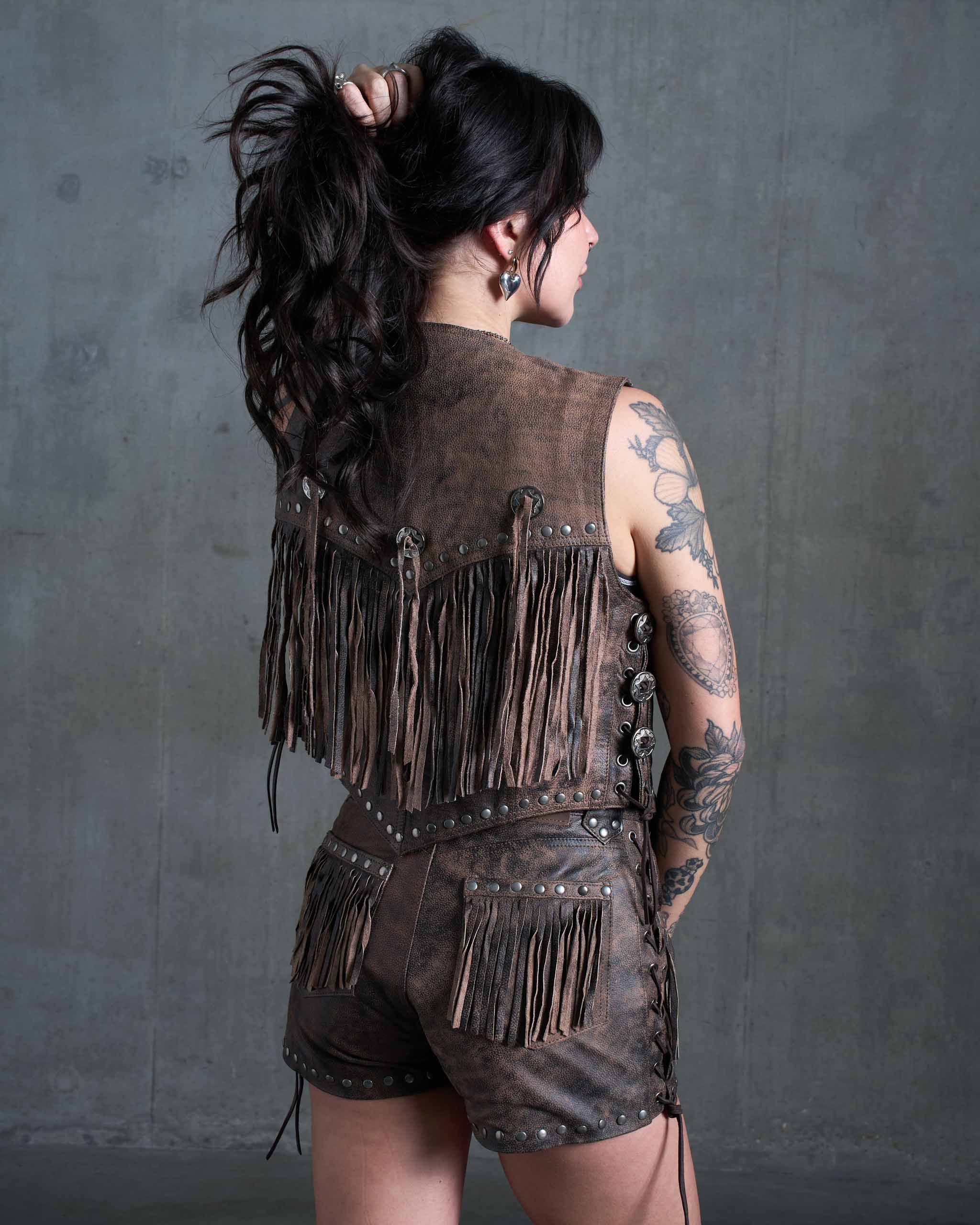 A tattooed model wearing the Sleazy Rider Colt Studded Fringe Leather vest and shorts set with a concrete backdrop.