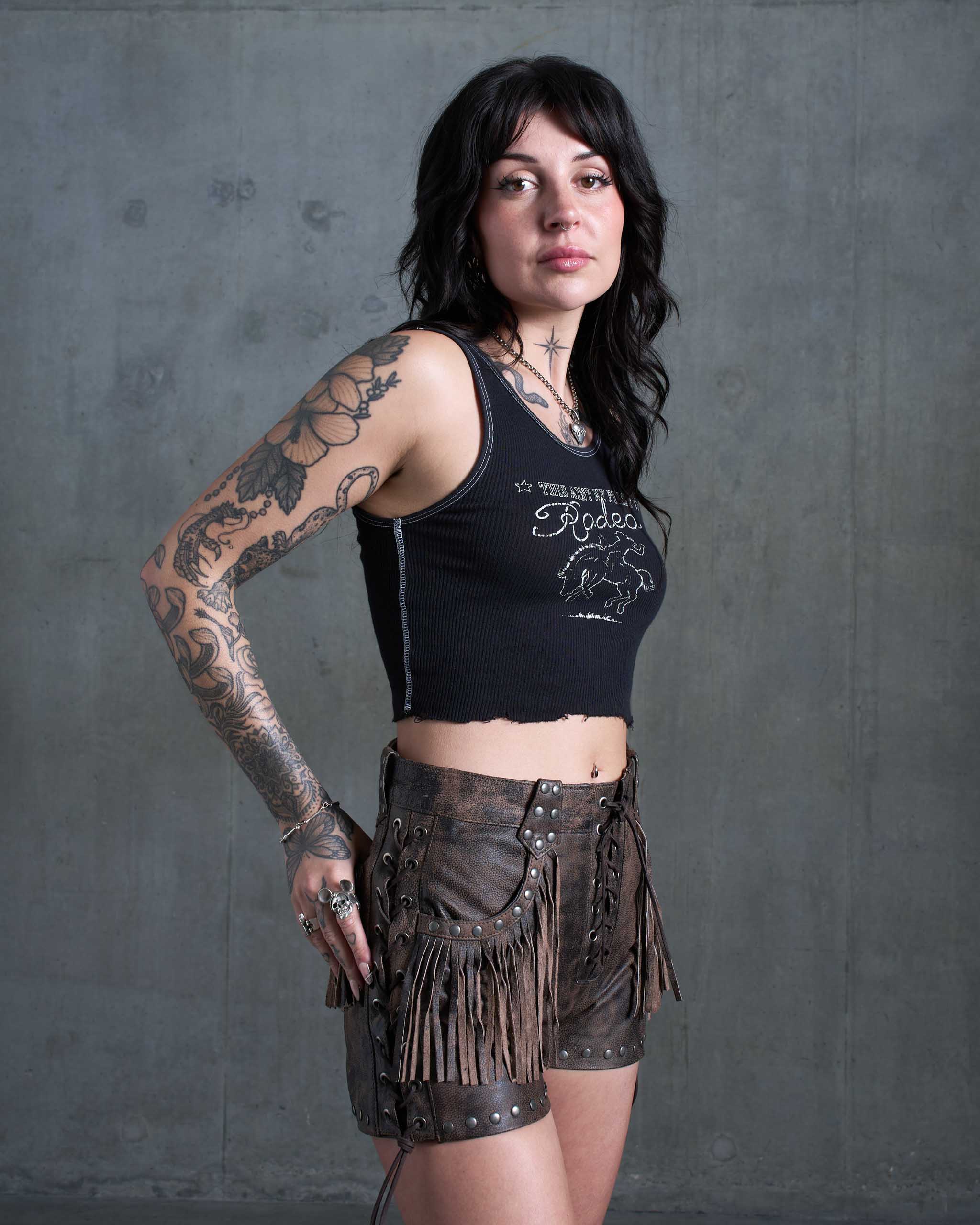A tattooed model wearing the Sleazy Rider Colt Studded Fringe Leather vest and shorts set with a concrete backdrop.