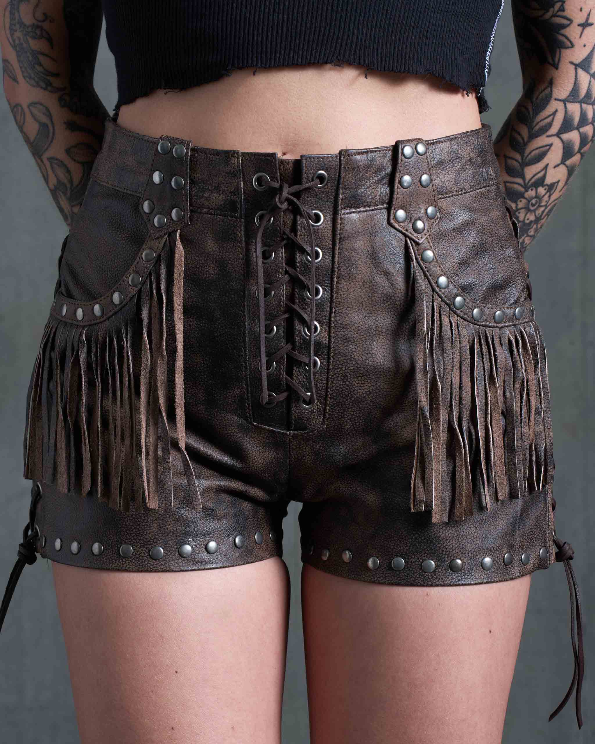A tattooed model wearing the Sleazy Rider Colt Studded Fringe Leather vest and shorts set with a concrete backdrop.