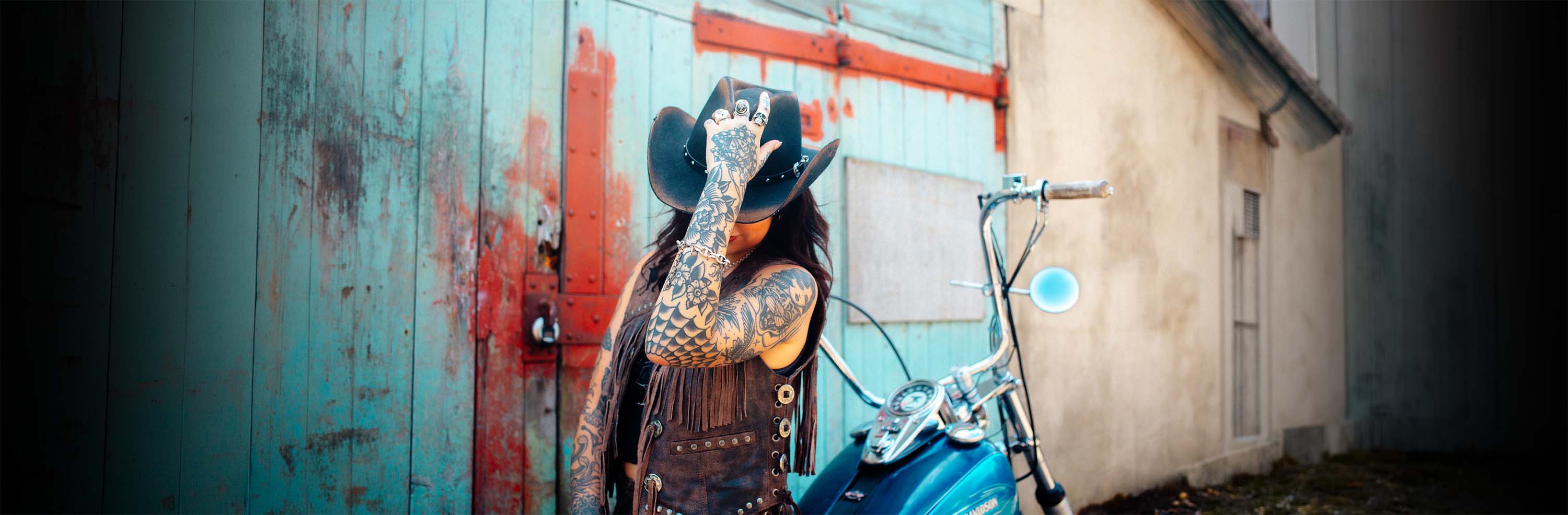 A tattooed cowgirl wearing a brown Sleazy Rider wild western leather vest.