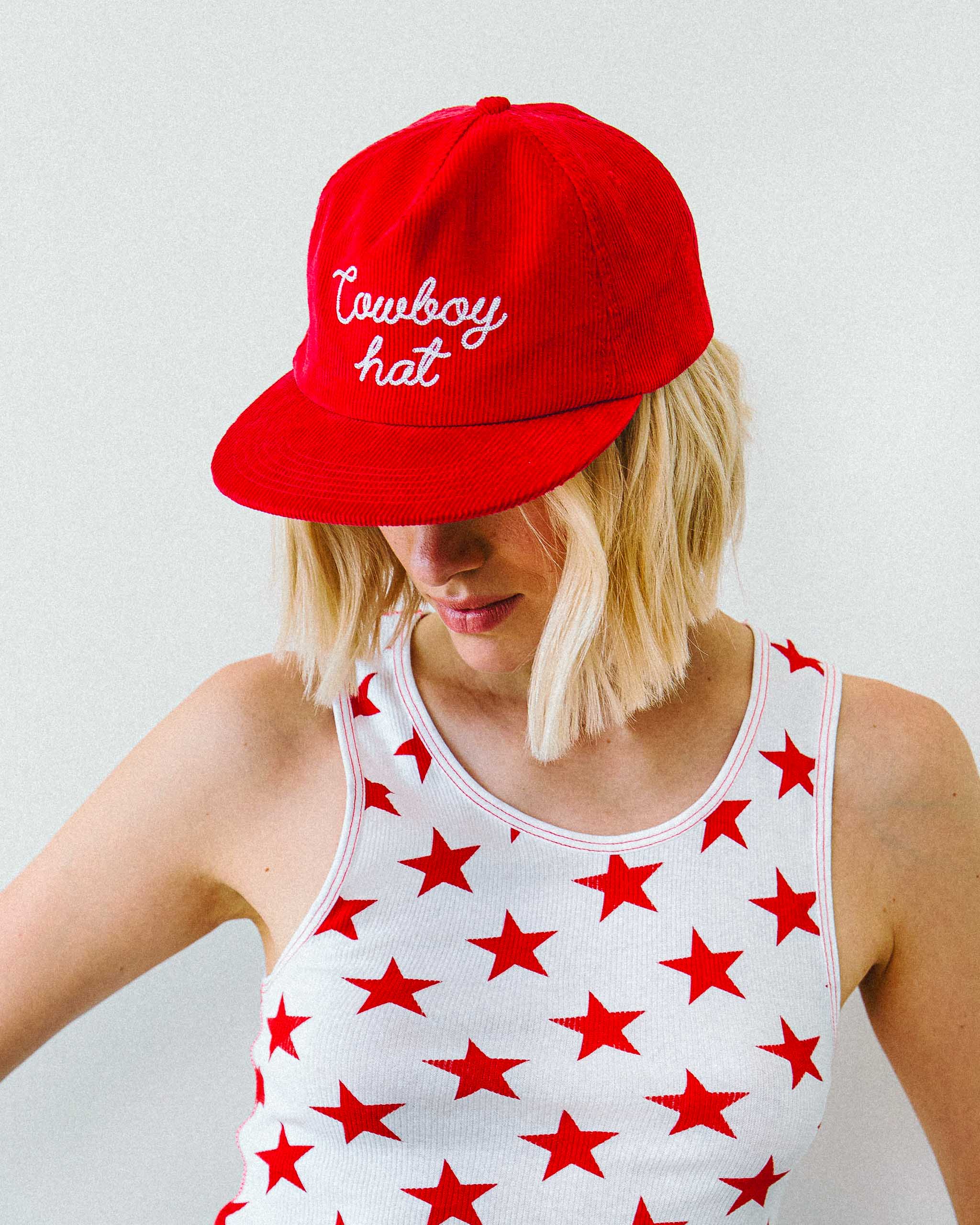 A model wearing a white tank top with red stars printed all over and a red cap with "Cowbot hat" embroidery.