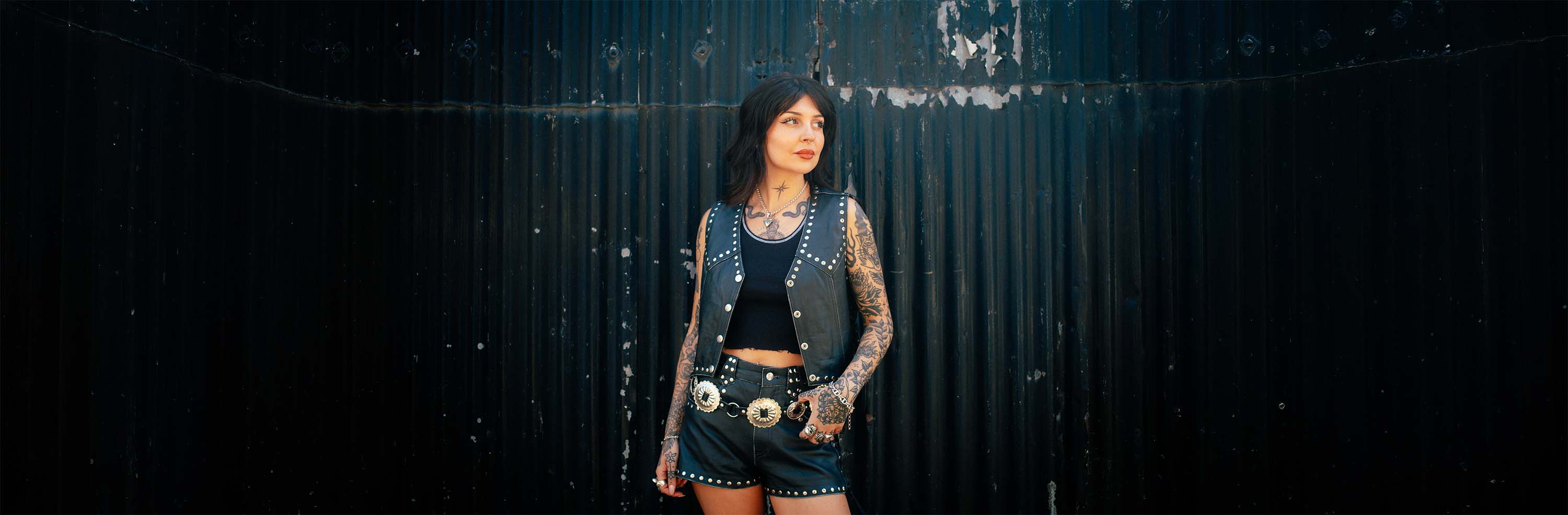 A biker woman standing in front of a black corrugated wall wearing Sleazy Rider coyote studded leather vest and shorts.