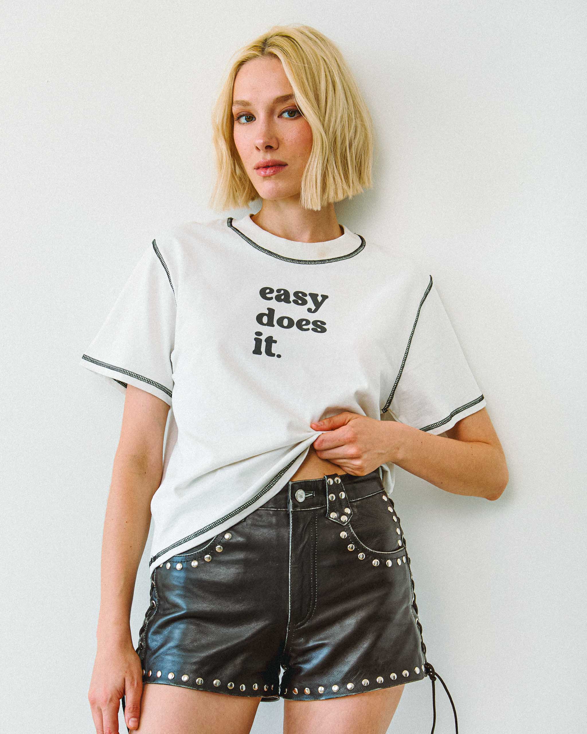 Woman wearing a white t-shirt with 'easy does it' text and black leather shorts against a plain background.
