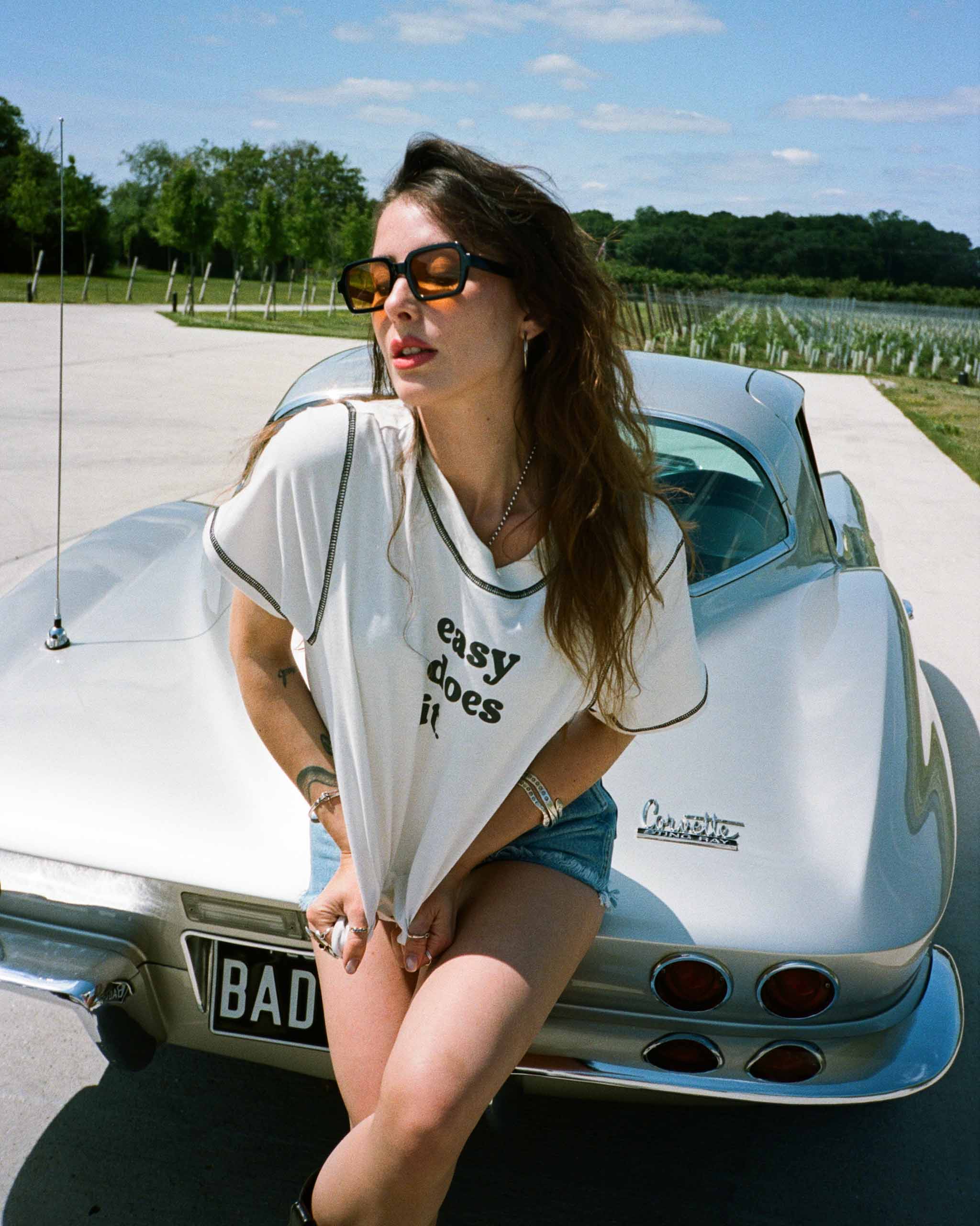 A young woman sitting on a classic car with a scenic background wearing a vintage style t-shirt that says 'Easy Does It".