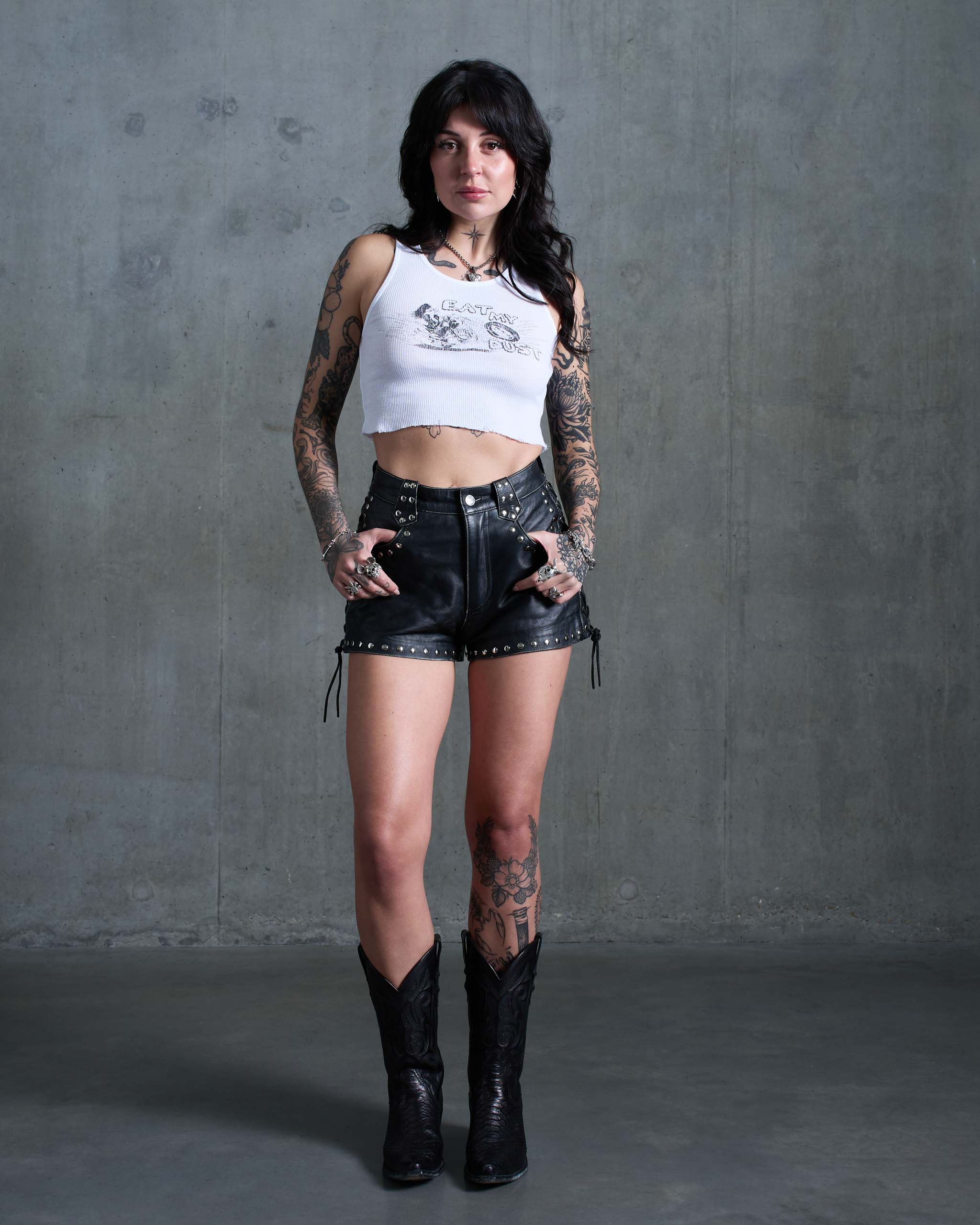 A tattooed woman wearing an 'Eat My dust' biker tank top, studded leather shorts, and black cowboy boots.