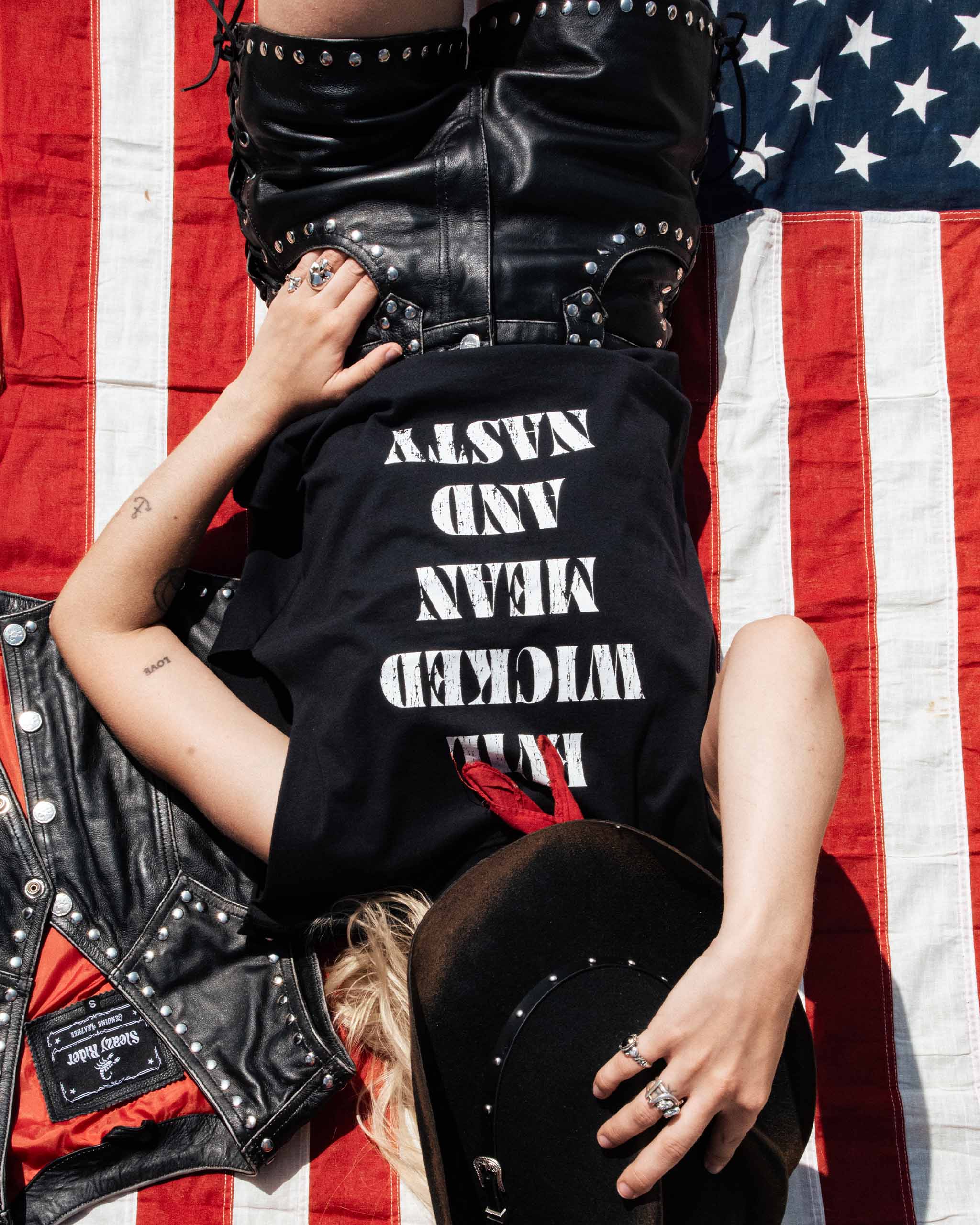 A female biker wearing a black cut off t-shirt with white text saying "Evil Wicked Mean and Nasty" on it, lying on an American flag.