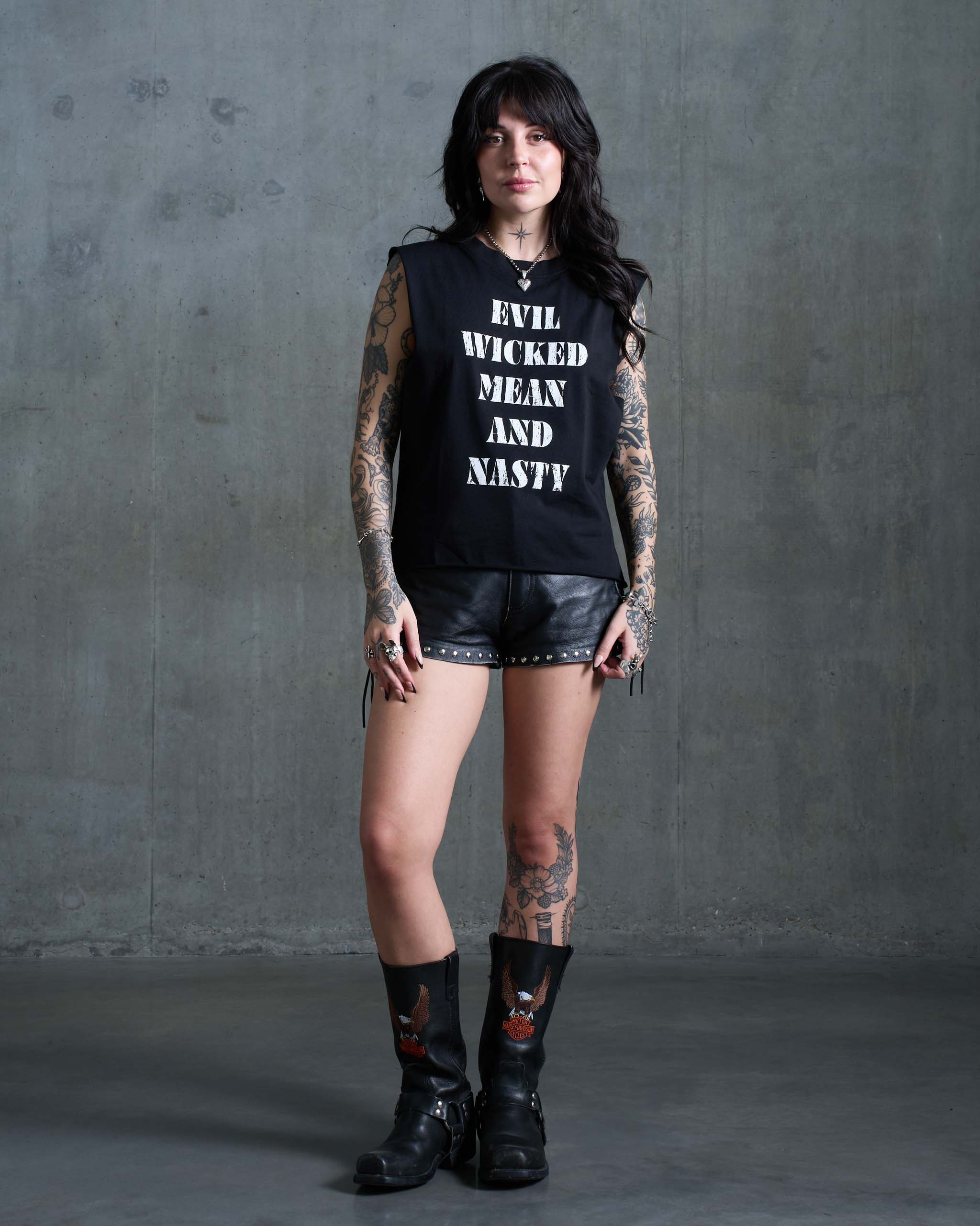 A biker chick wearing an 'EVIL WICKED MEAN AND NASTY' t-shirt with cut-off sleeves, studded leather shorts, and black cowboy boots.
