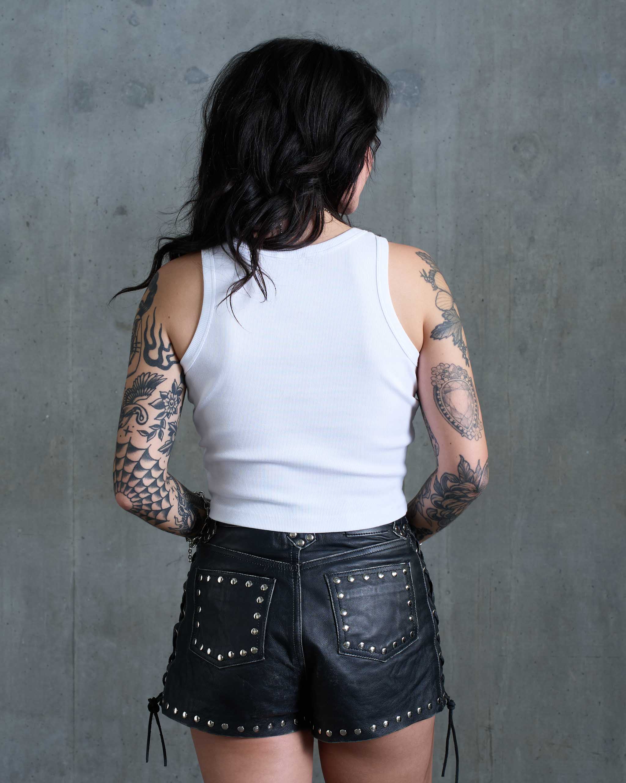 A tattooed woman wearing a white tank top and studded leather shorts, view from behind.