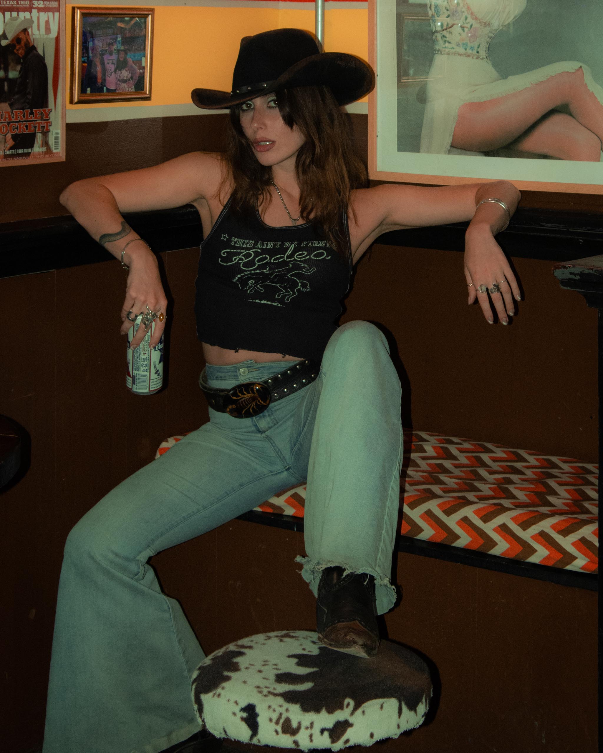 Woman in cowboy hat and rodeo tank top, sitting on a couch with a drink.