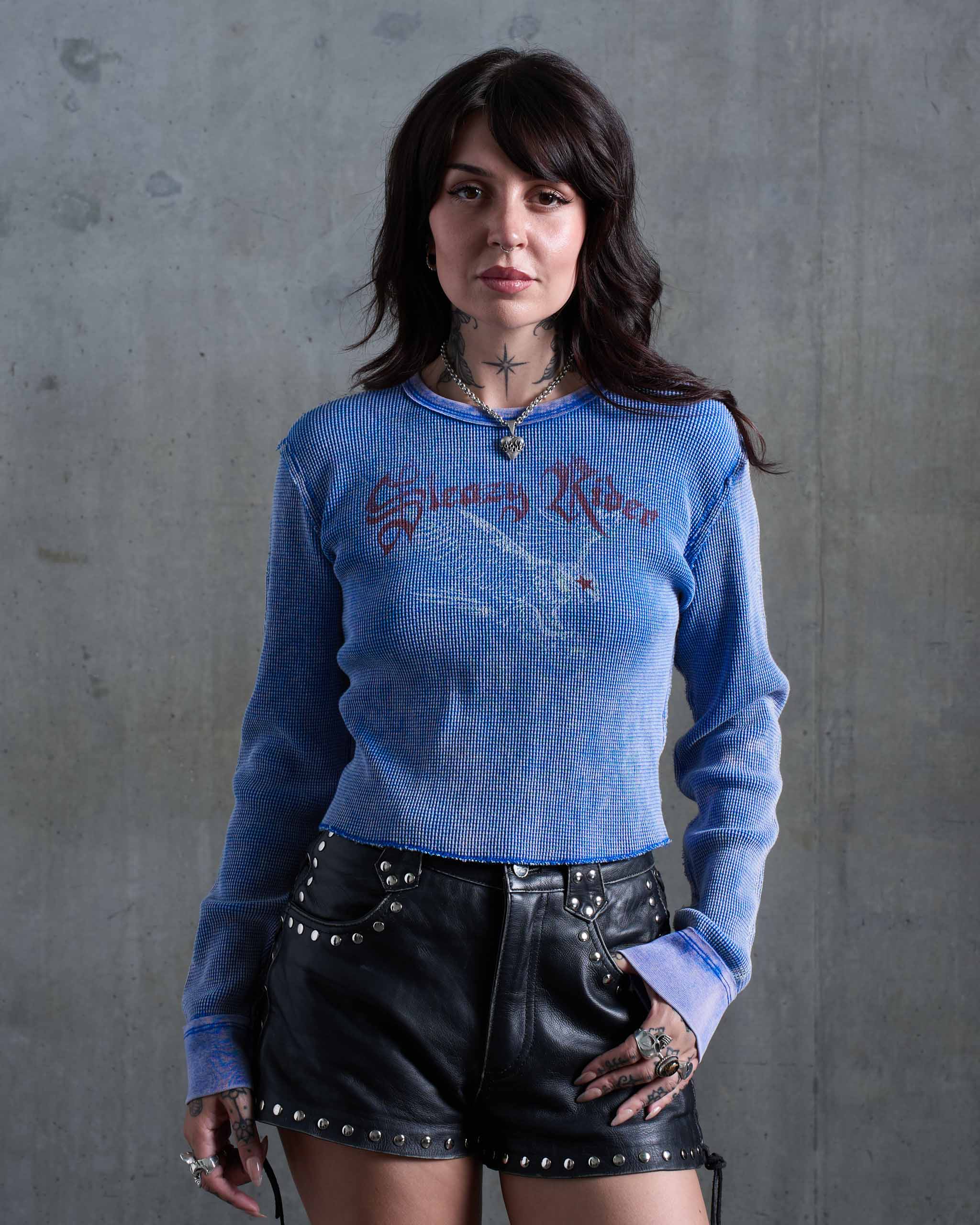 A tattooed model wearing a blue faded vintage waffle knit top with a swooping eagle printed on the front, with black studded leather shorts.