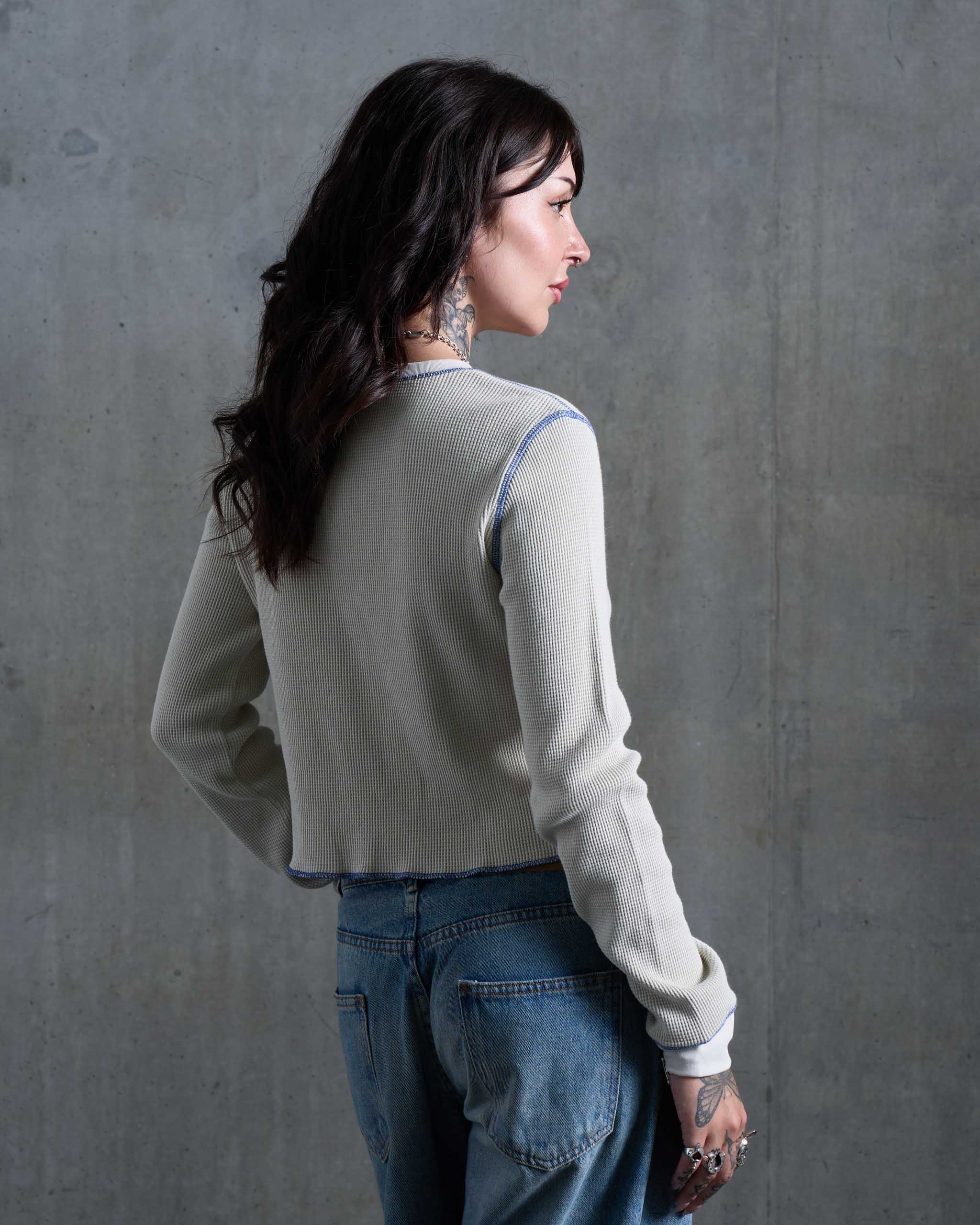 A tattooed woman wearing an off-white waffle knit top, with low rise jeans, viewed from behind.