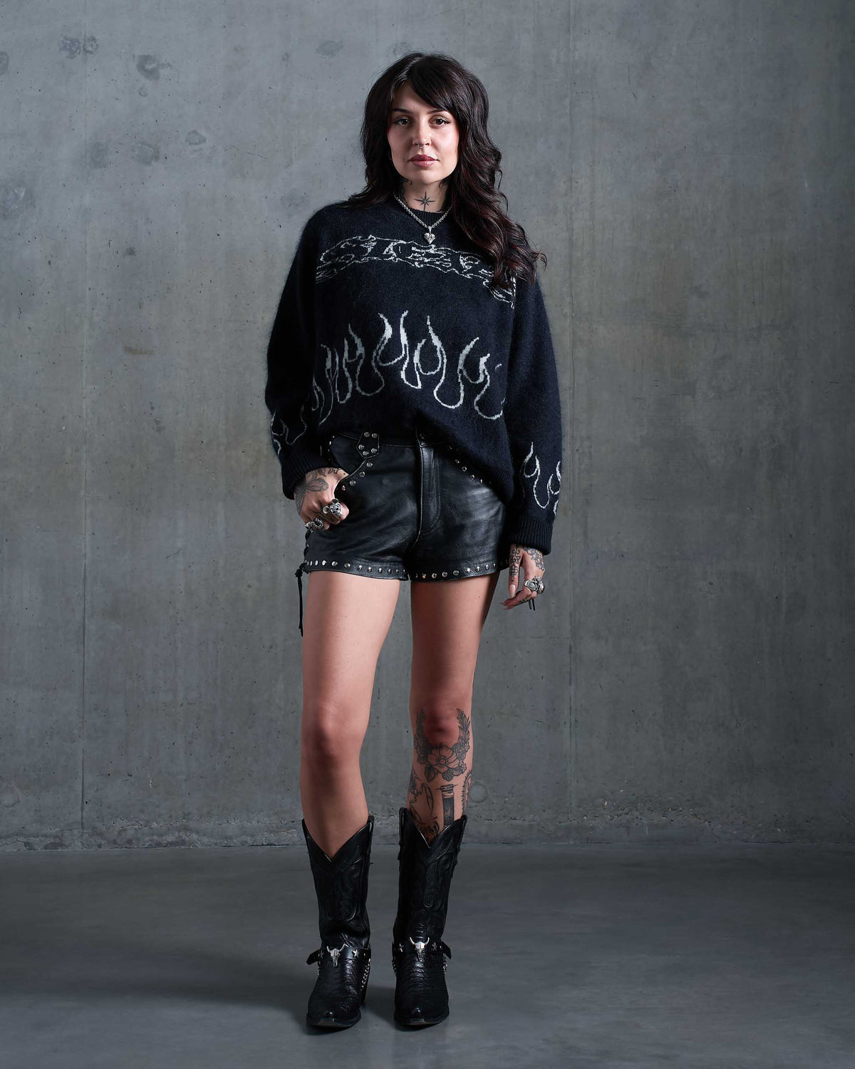 An alt girl with tattoos wearing a gothic black knitted jumper with white flames, studded leather shorts, and cowboy boots. Standing in an industrial concrete room.