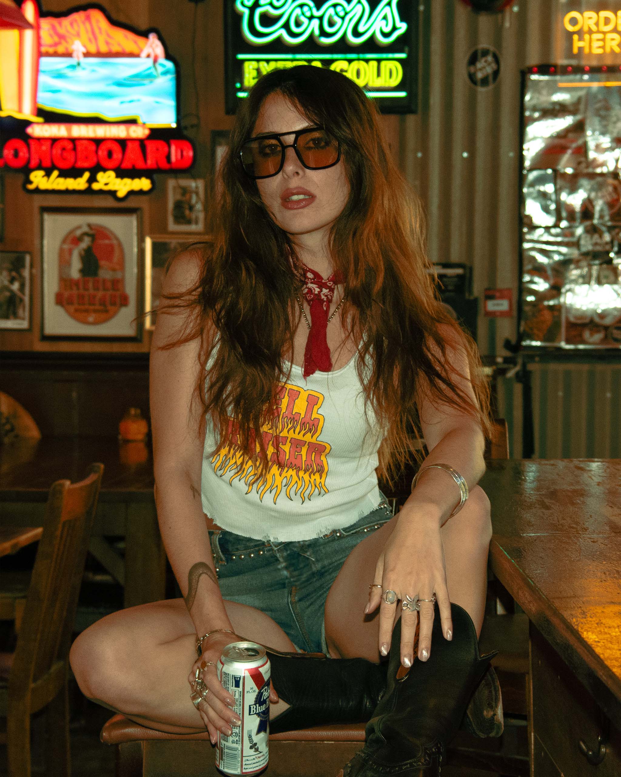 A woman sitting at an American dive bar, wearing a 'Hell Raiser' tank top.