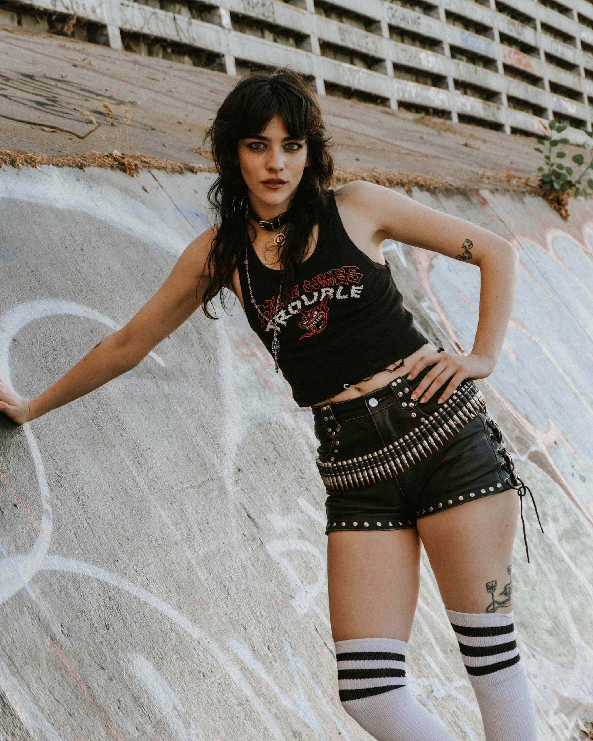 An alternative model wearing a "Here Comes Trouble" black tank top with studded leather shorts and knee high socks against a concrete backdrop.