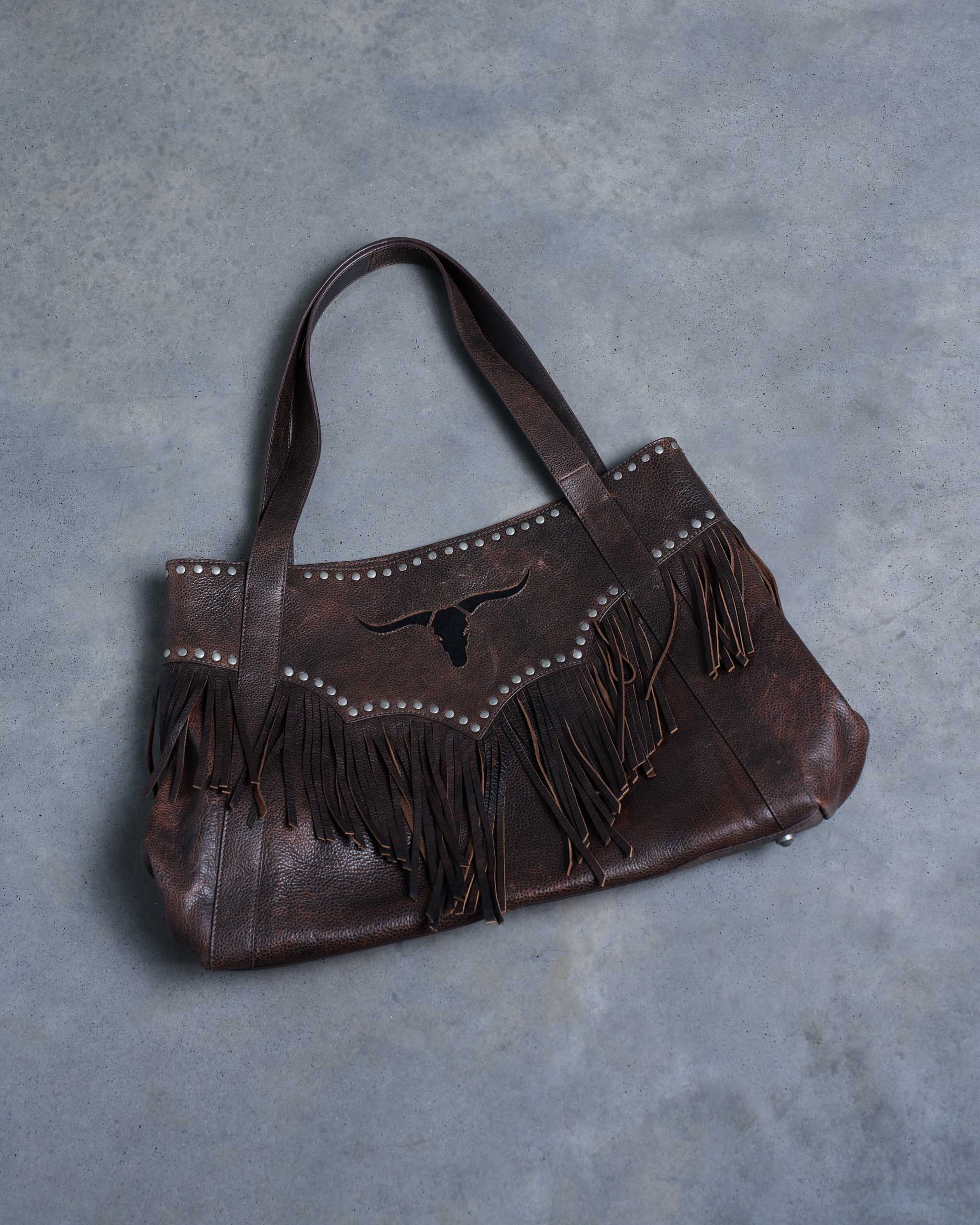 A large wild western bag made from brown distressed leather, with studs, fringe and a black suede texas longhorn emblem on the front.