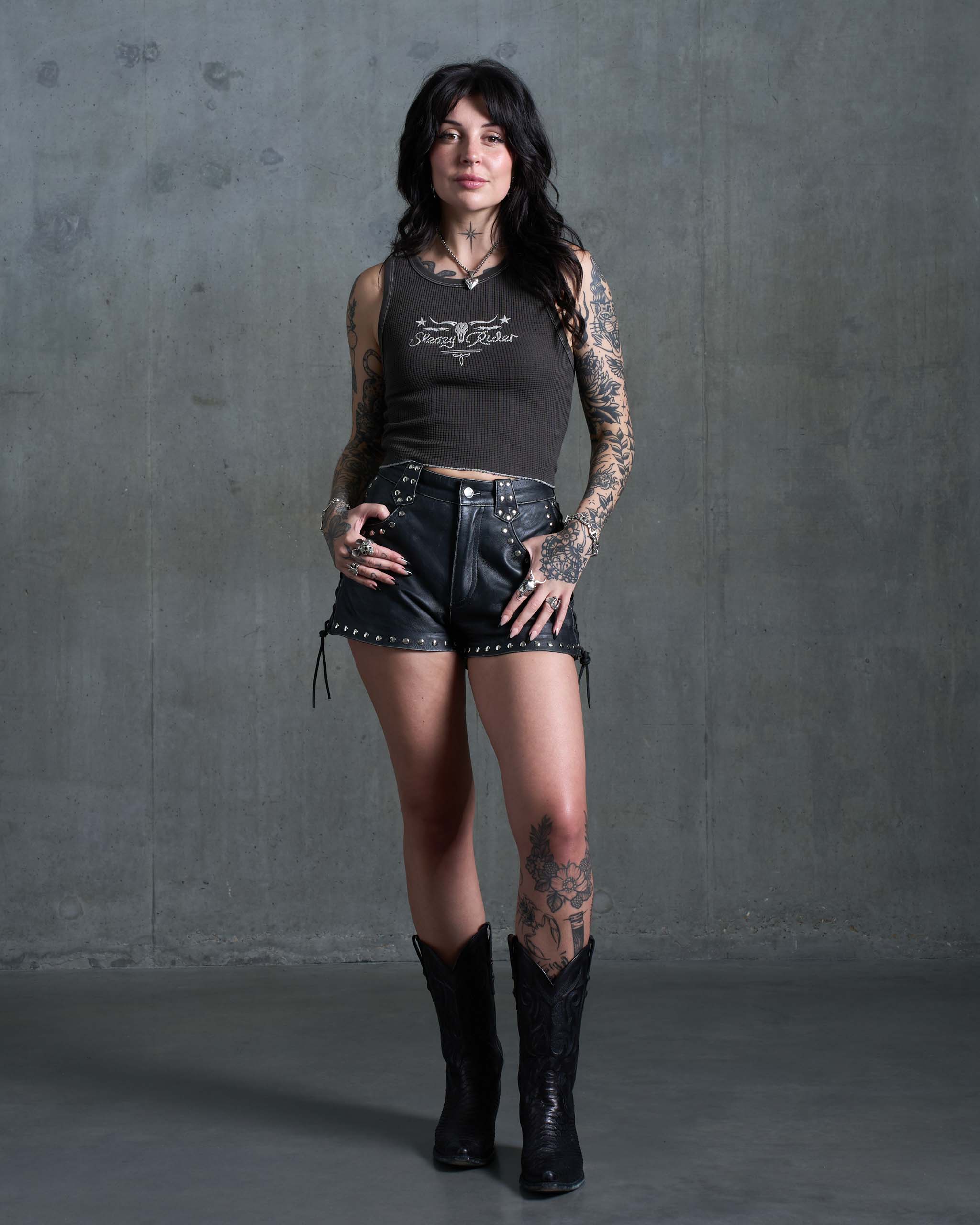 A gothic western woman wearing a waffle knit tank top with a longhorn skull embroidery, studded leather shorts and cowboy boots.