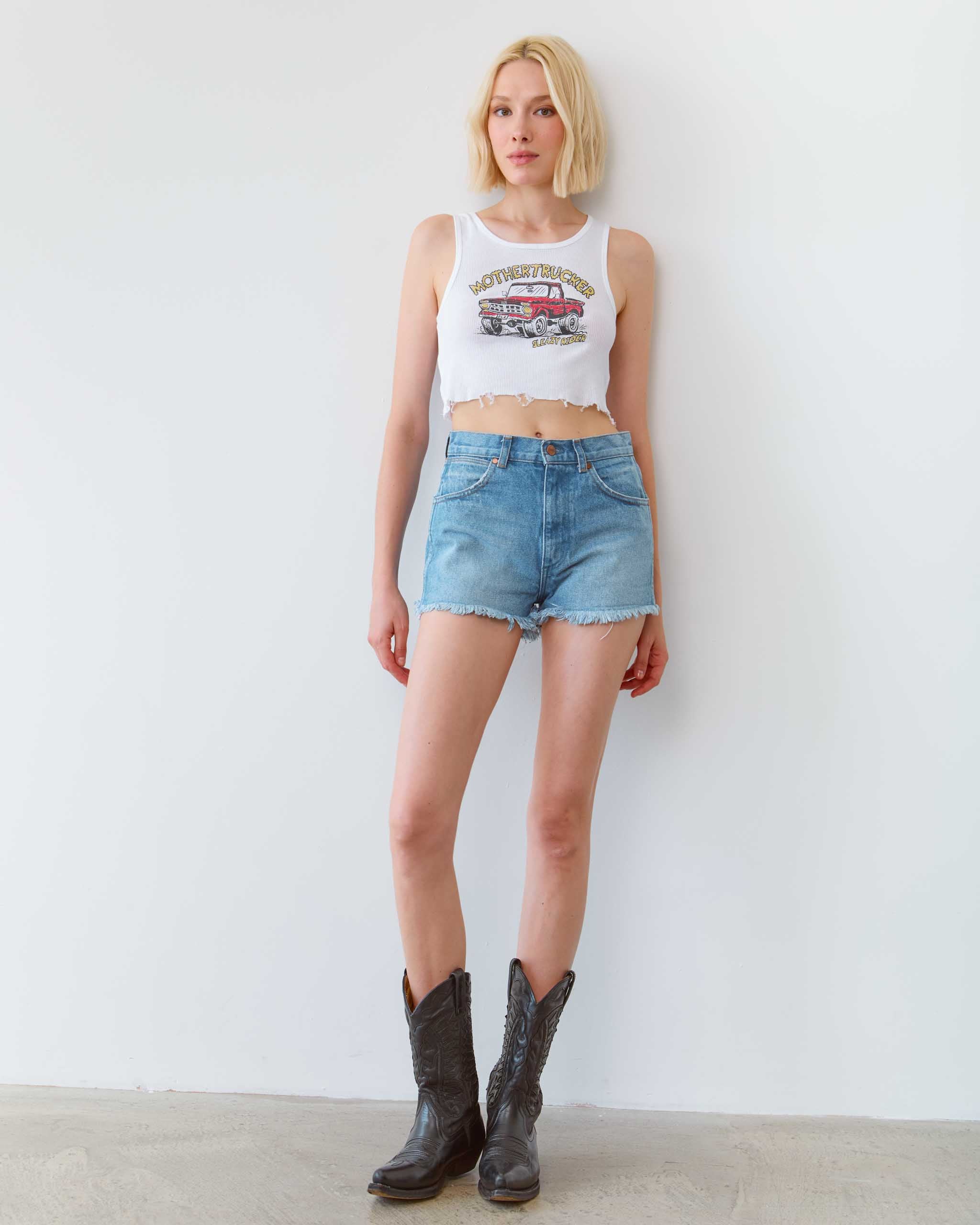 Woman wearing a white tank top with a graphic design, denim shorts, and brown boots against a white background
