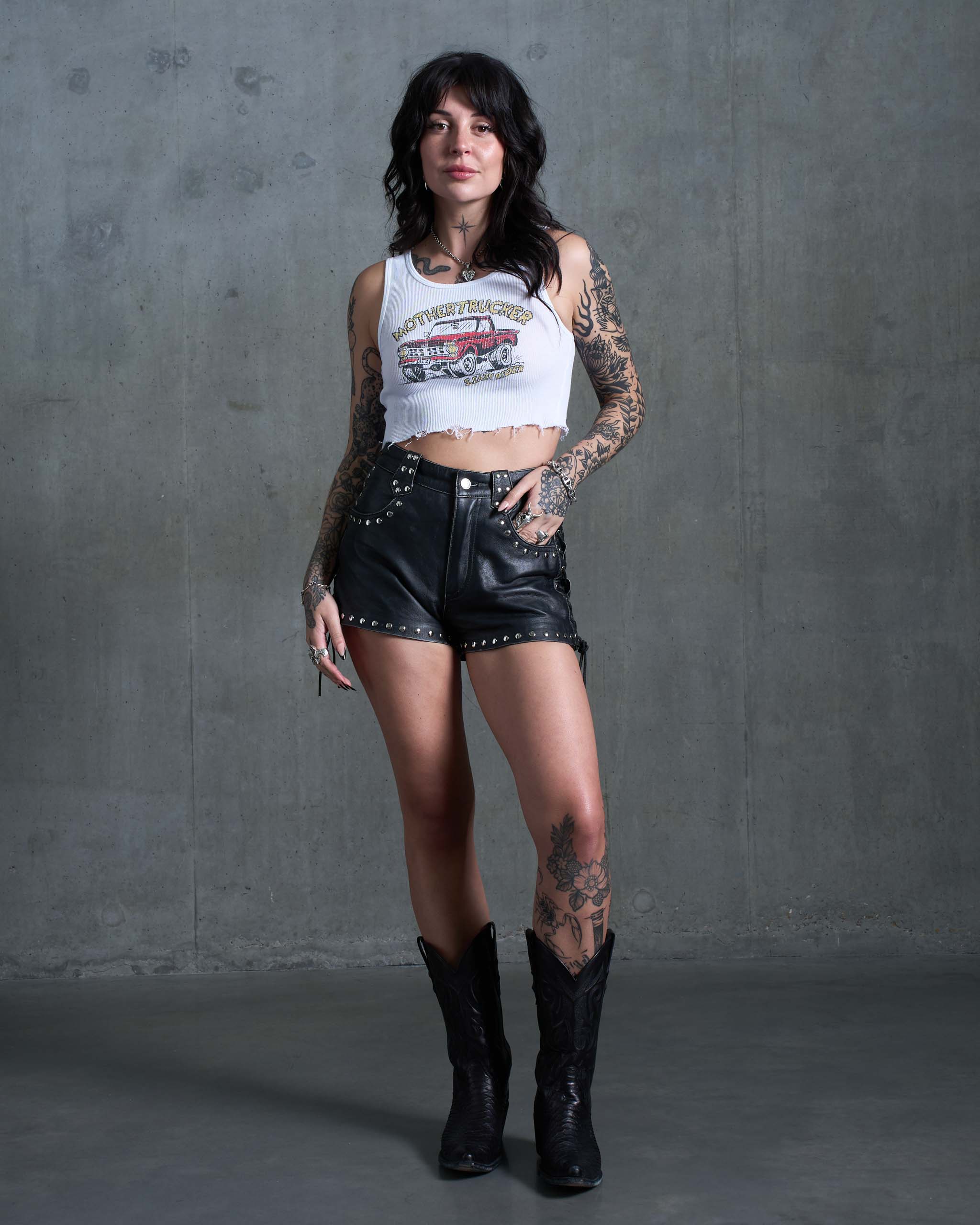 A tattooed model wearing a vintage truck tank top, studded leather shorts and cowboy boots.