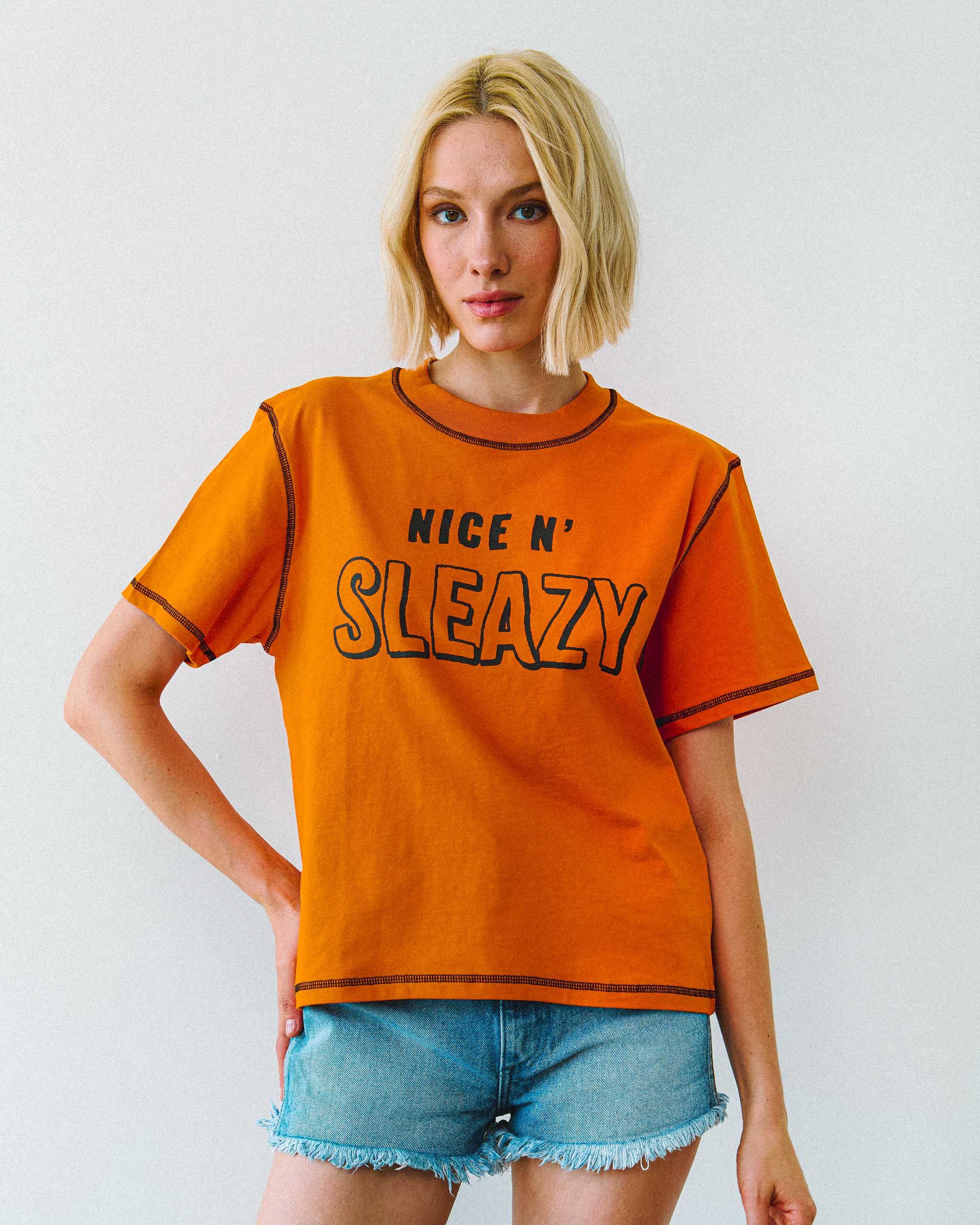 A young model wearing an orange "NICE N' SLEAZY" slogan t-shirt with denim shorts.