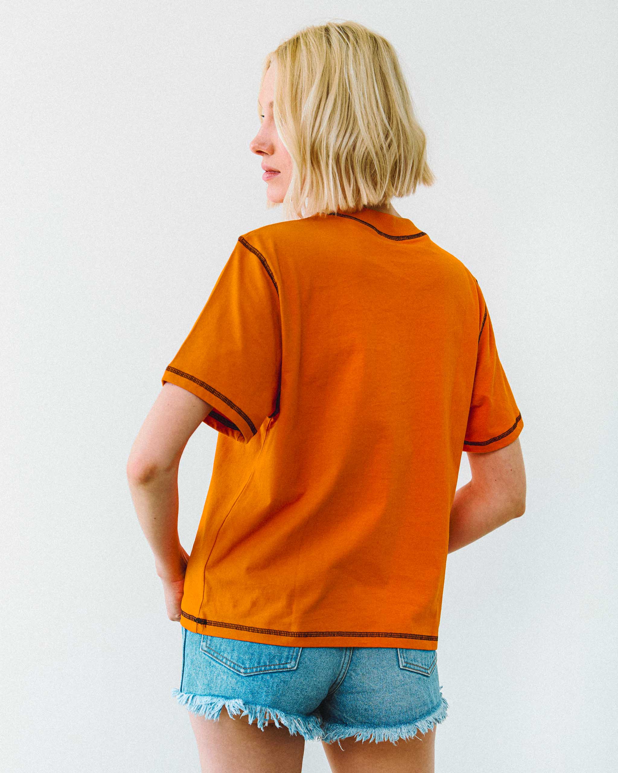 Person wearing an orange t-shirt shirt with black contrast stitching on a white background.