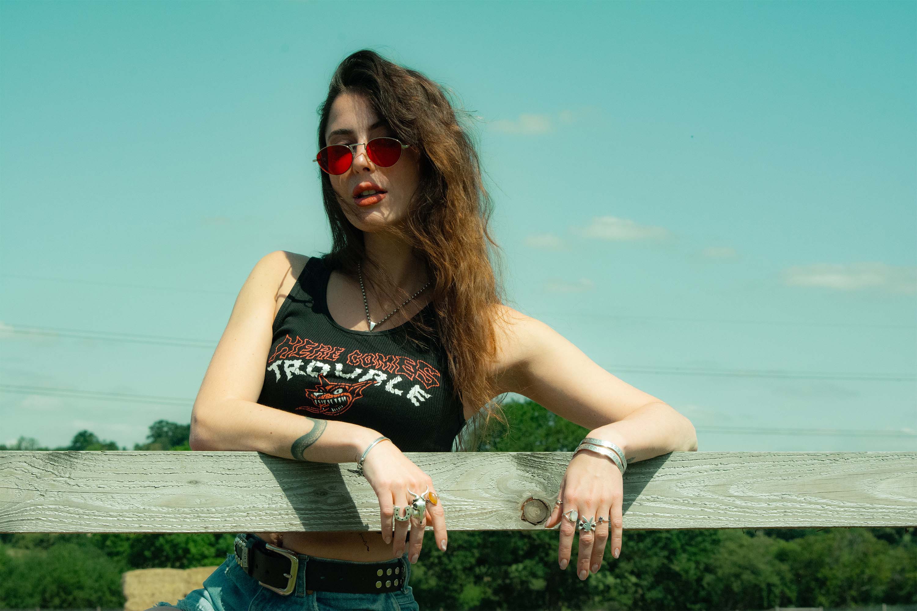 Woman wearing sunglasses and a tank top, leaning on a wooden fence with a clear sky background at woodstock.