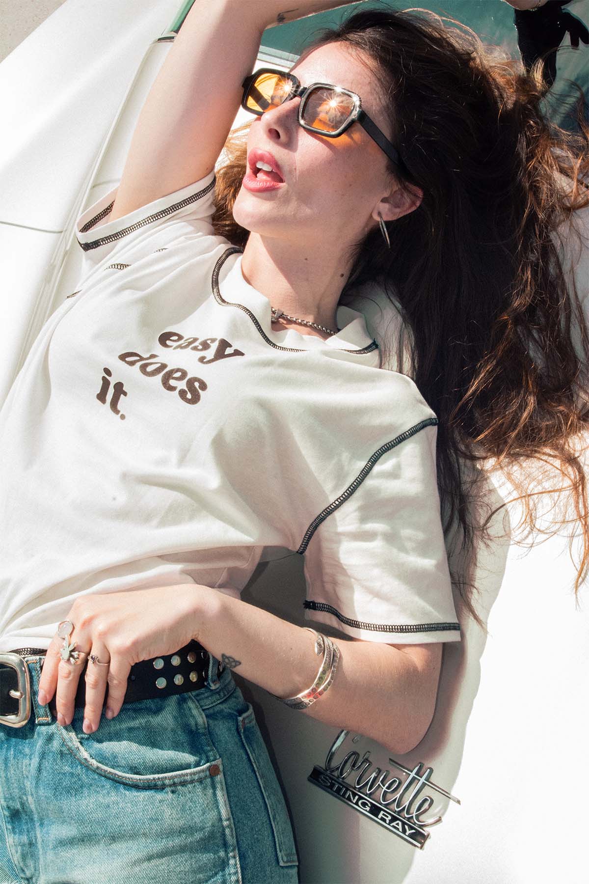 Woman wearing a white shirt with 'easy does it' text, blue jeans, and sunglasses, lying on a classic car.
