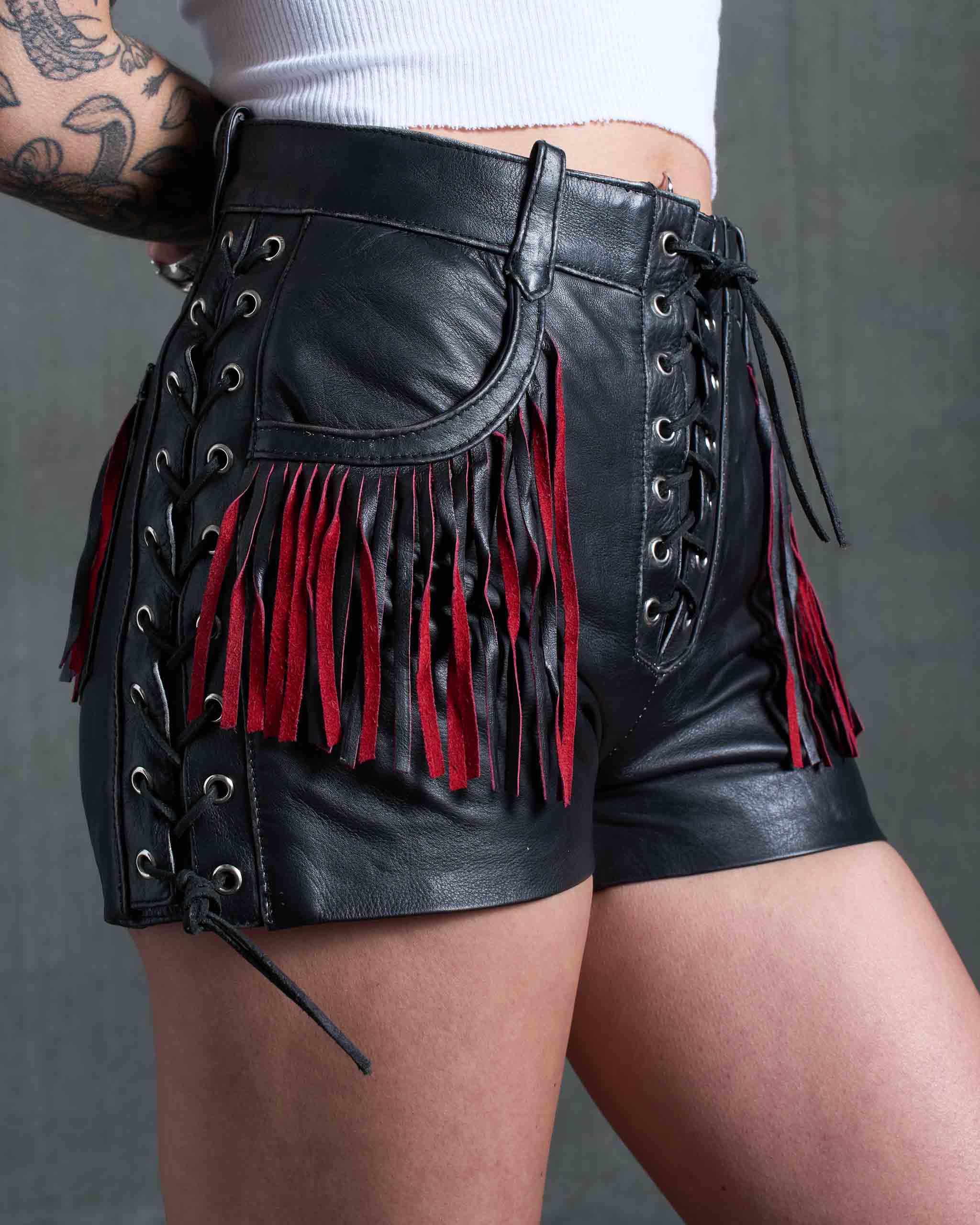A tattooed female model wearing the Sleazy Rider Outlaw Fringe Biker Leather Shorts, with lace-up and fringe.