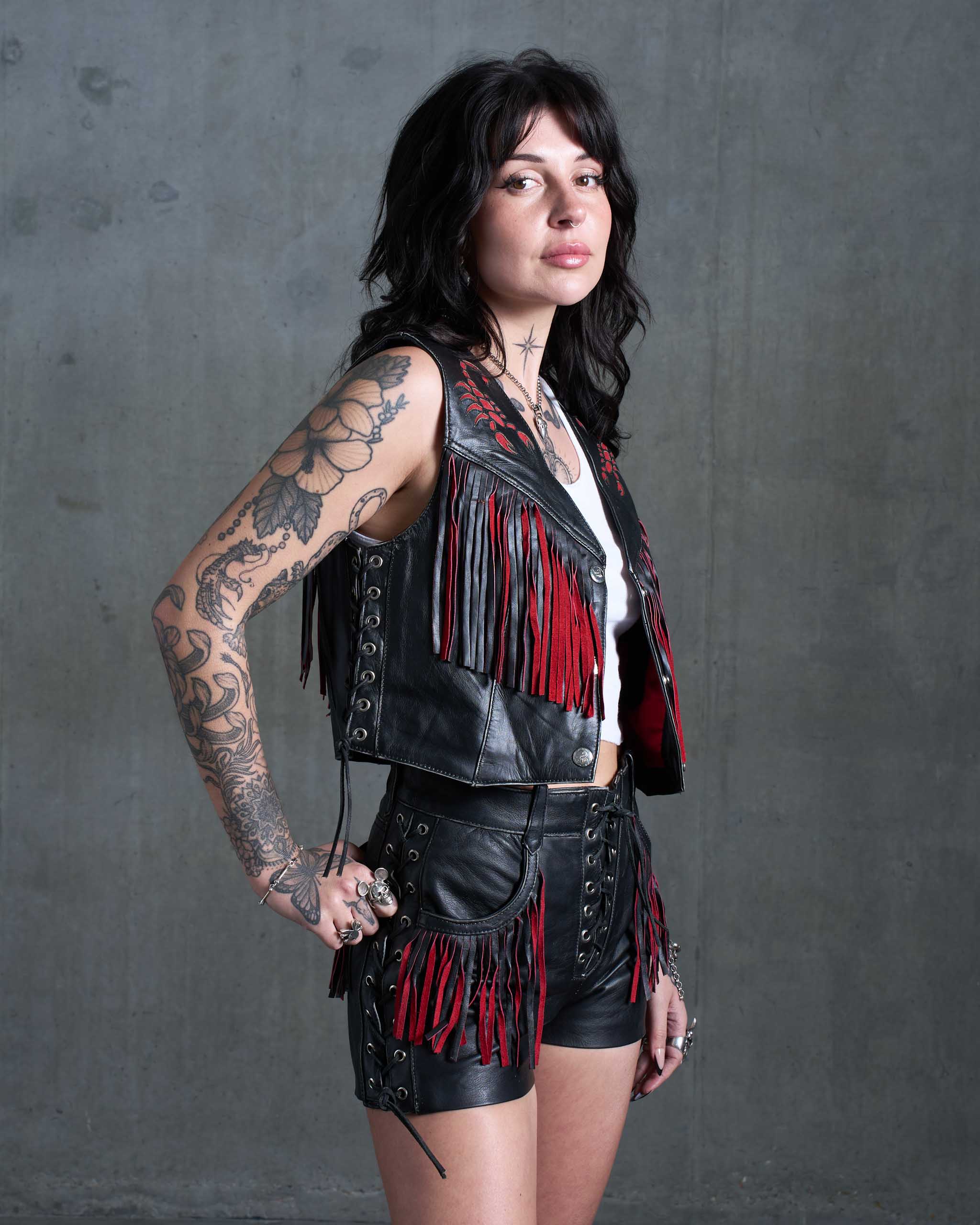 A tattooed model wearing the Sleazy Rider Outlaw Fringe Leather vest and shorts set with a concrete backdrop.