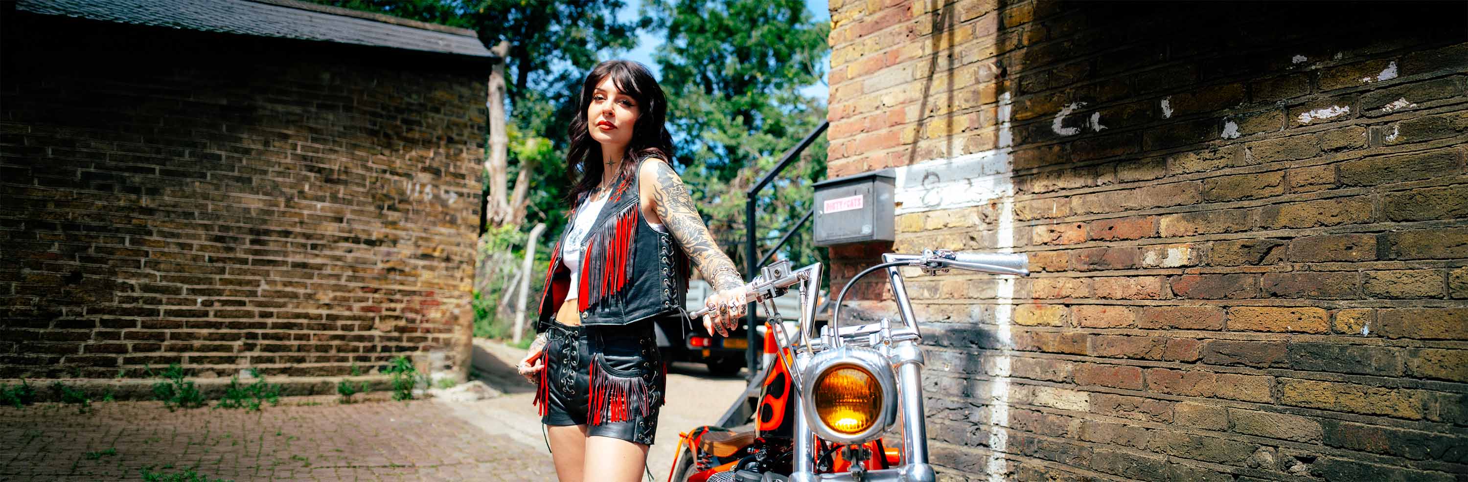 A tattooed biker woman standing with a custom chopper wearing black Sleazy Rider leather vest and shorts with red suede details.