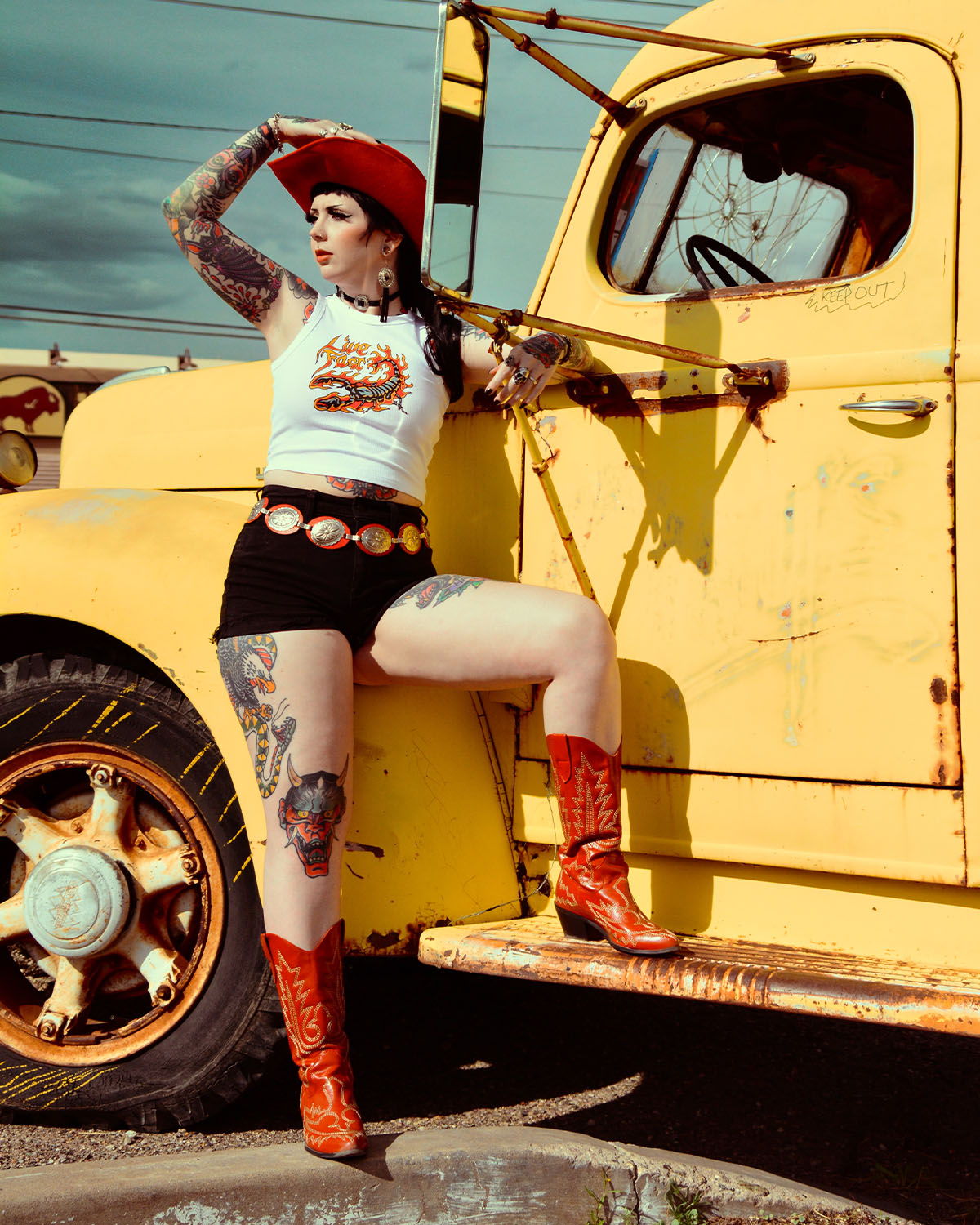 A gothic Western woman in a red cowboy hat and boots leaning against an old yellow truck.