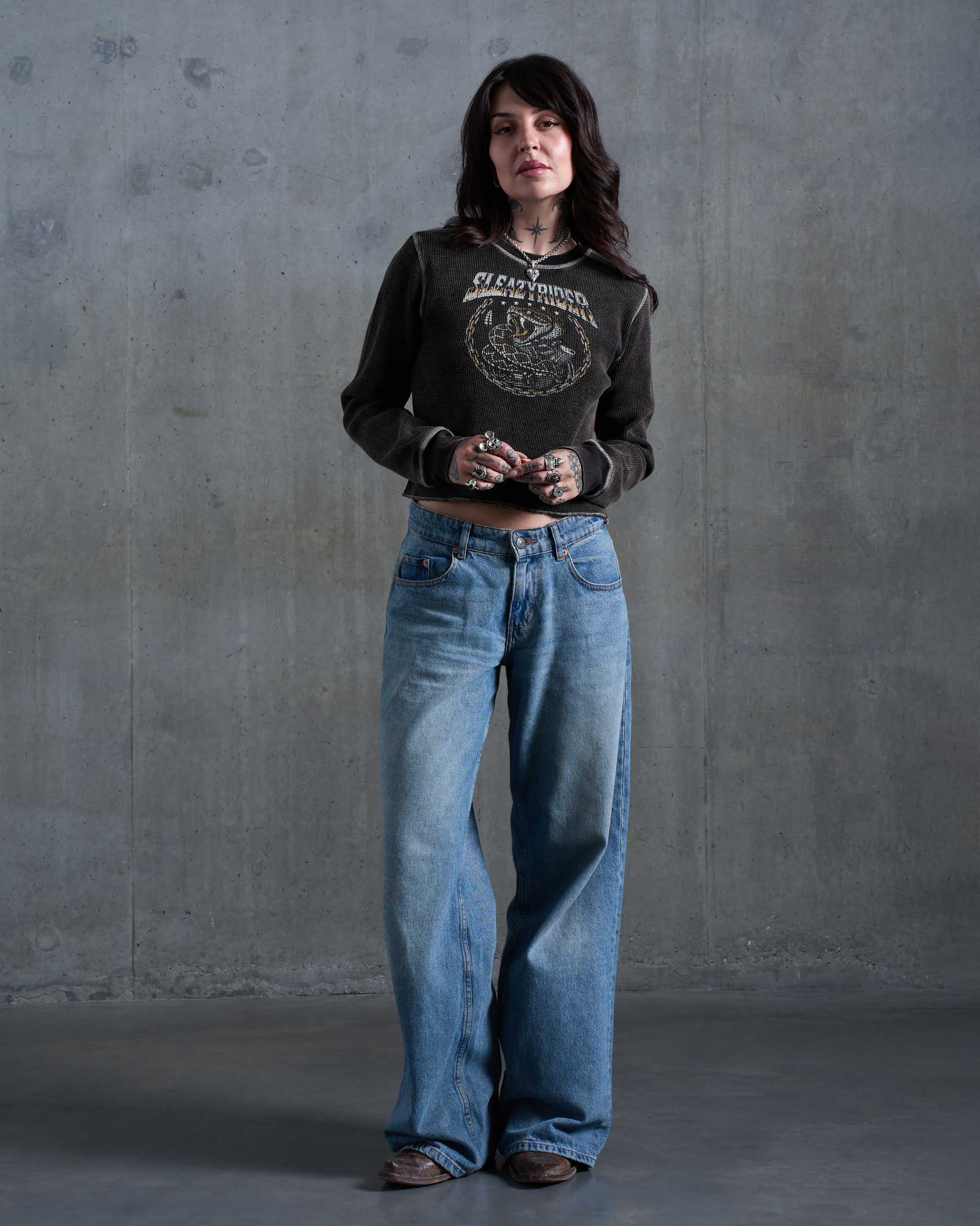 A tattooed woman wearing a vintage faded waffle knit top with a rattlesnake print, with low rise jeans and brown cowboy boots.