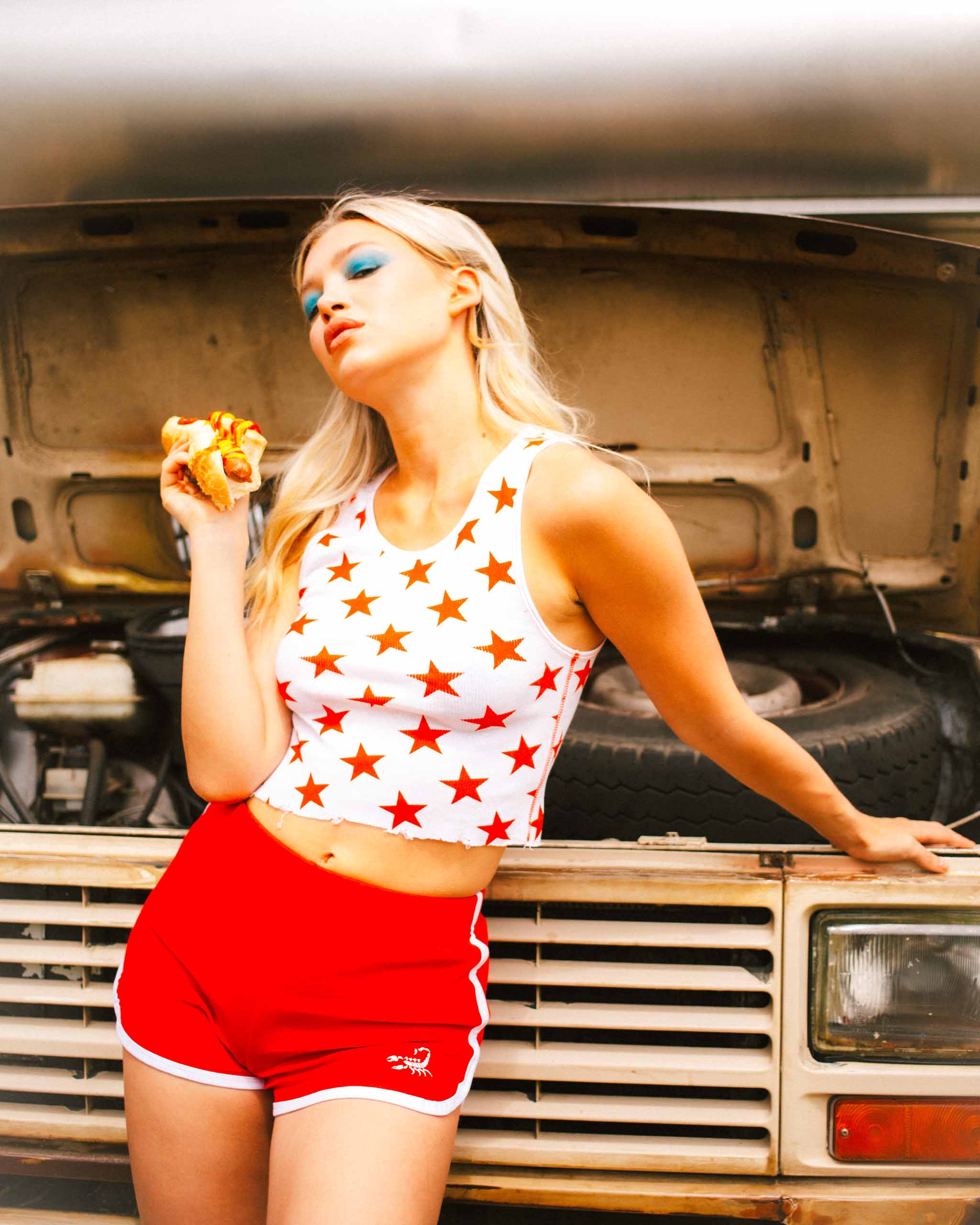 Woman in a white and red star print top and vintage style sport shorts standing in front of an open car hood eating a hotdog.