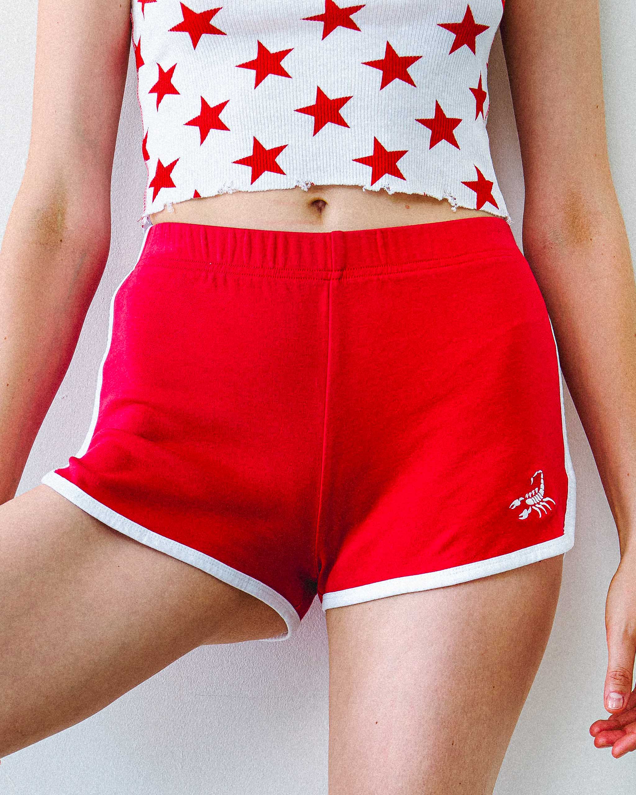 Person wearing a white top with red star pattern and red Sleazy Rider 70s shorts with scorpion embroidery on a plain background.