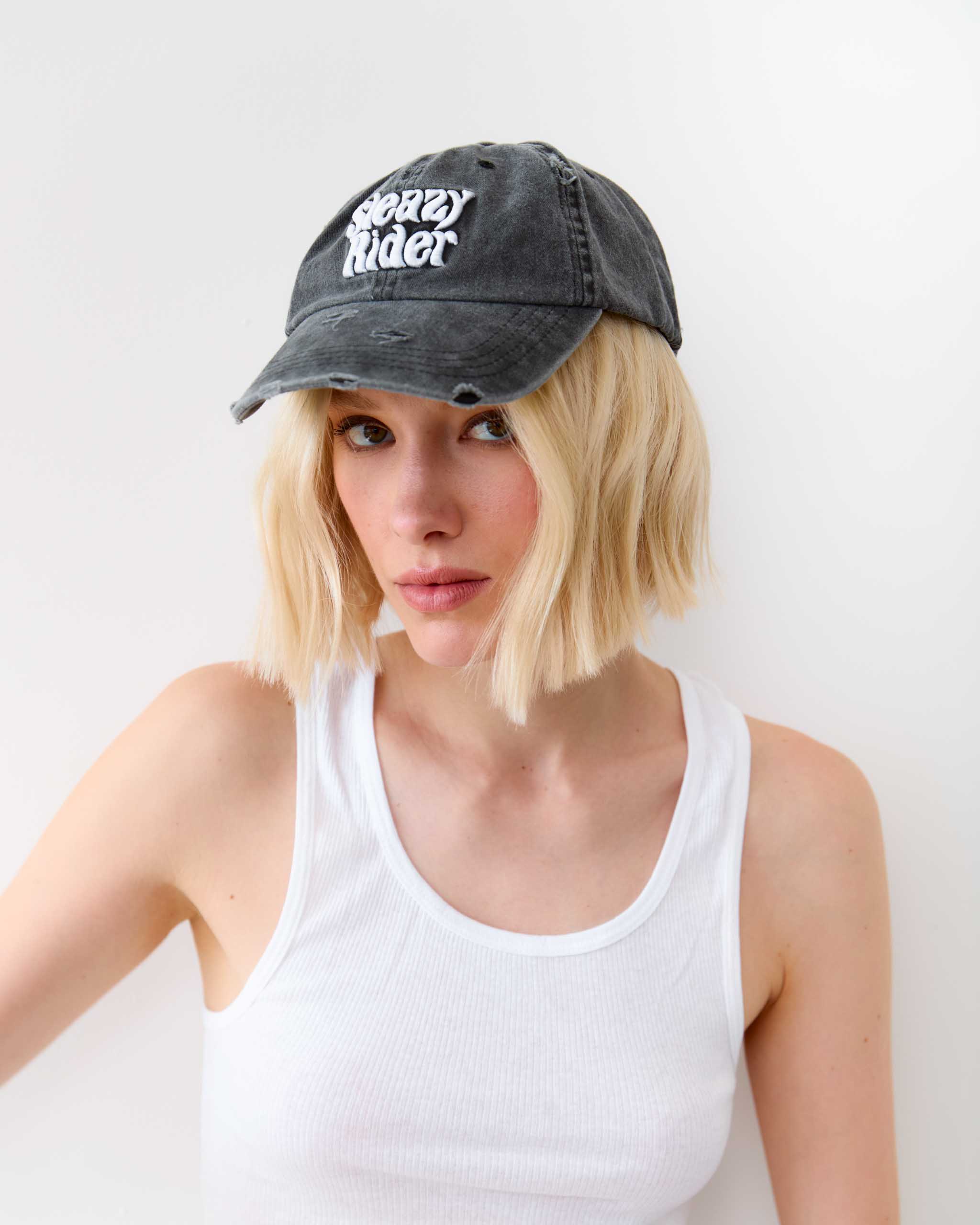 Woman wearing a distressed denim cap with 'Sleazy Rider" text on a white background